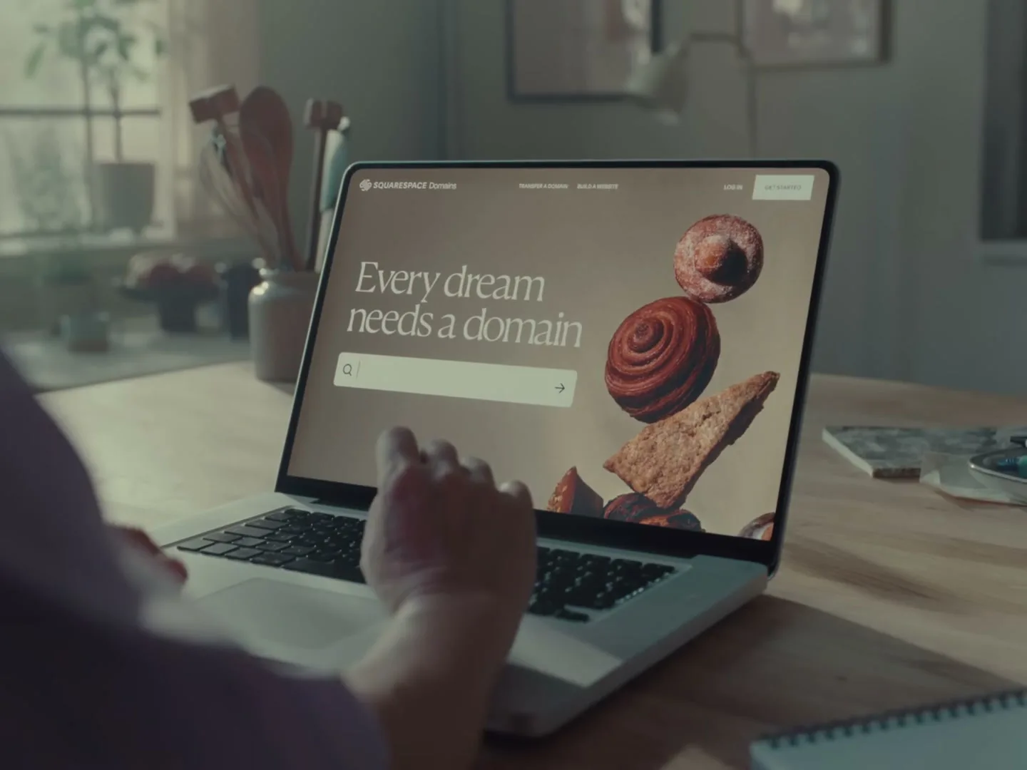 A person's hands type on a laptop displaying the Squarespace website with the text "Every dream needs a domain" in a sunlit kitchen.