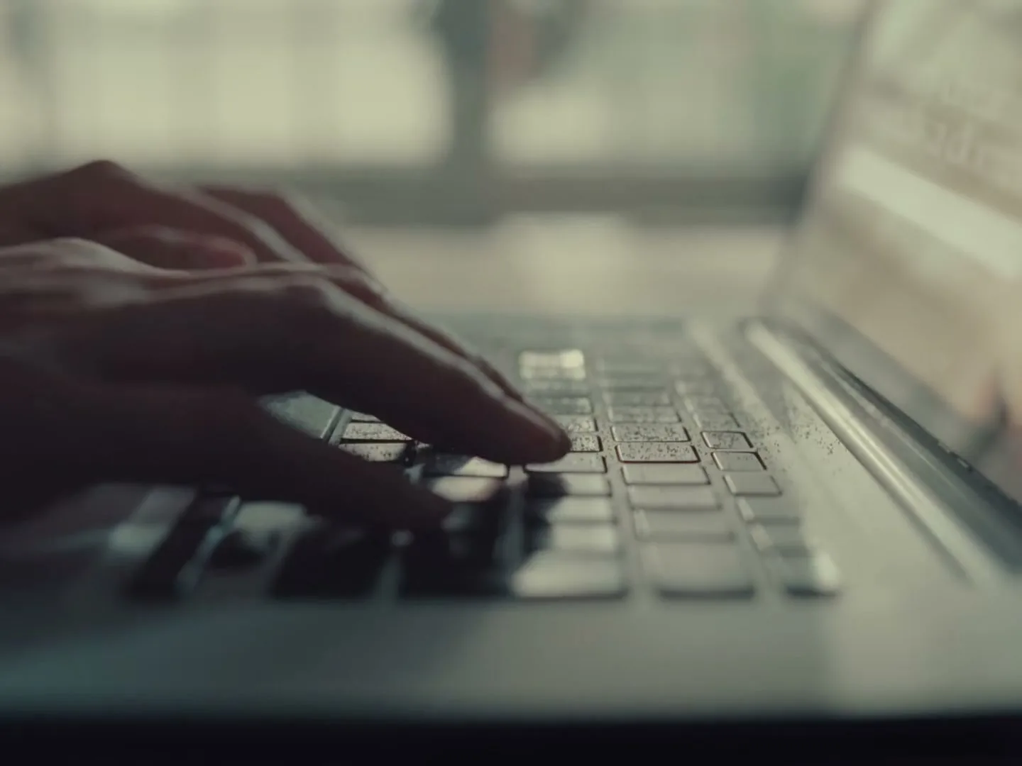 A person's hands are typing on a laptop keyboard, with a window and blurred background visible behind them.