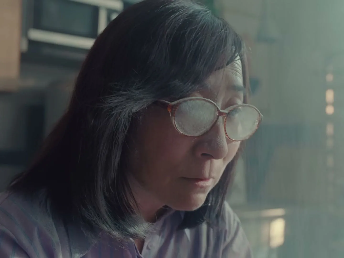 A woman with flour-dusted glasses looks down, concentrating on her baking in a kitchen.
