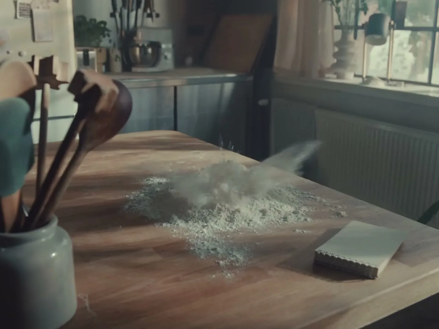 A pile of flour on a wooden kitchen counter is hit by an egg, causing a small explosion of flour in a sunlit kitchen.