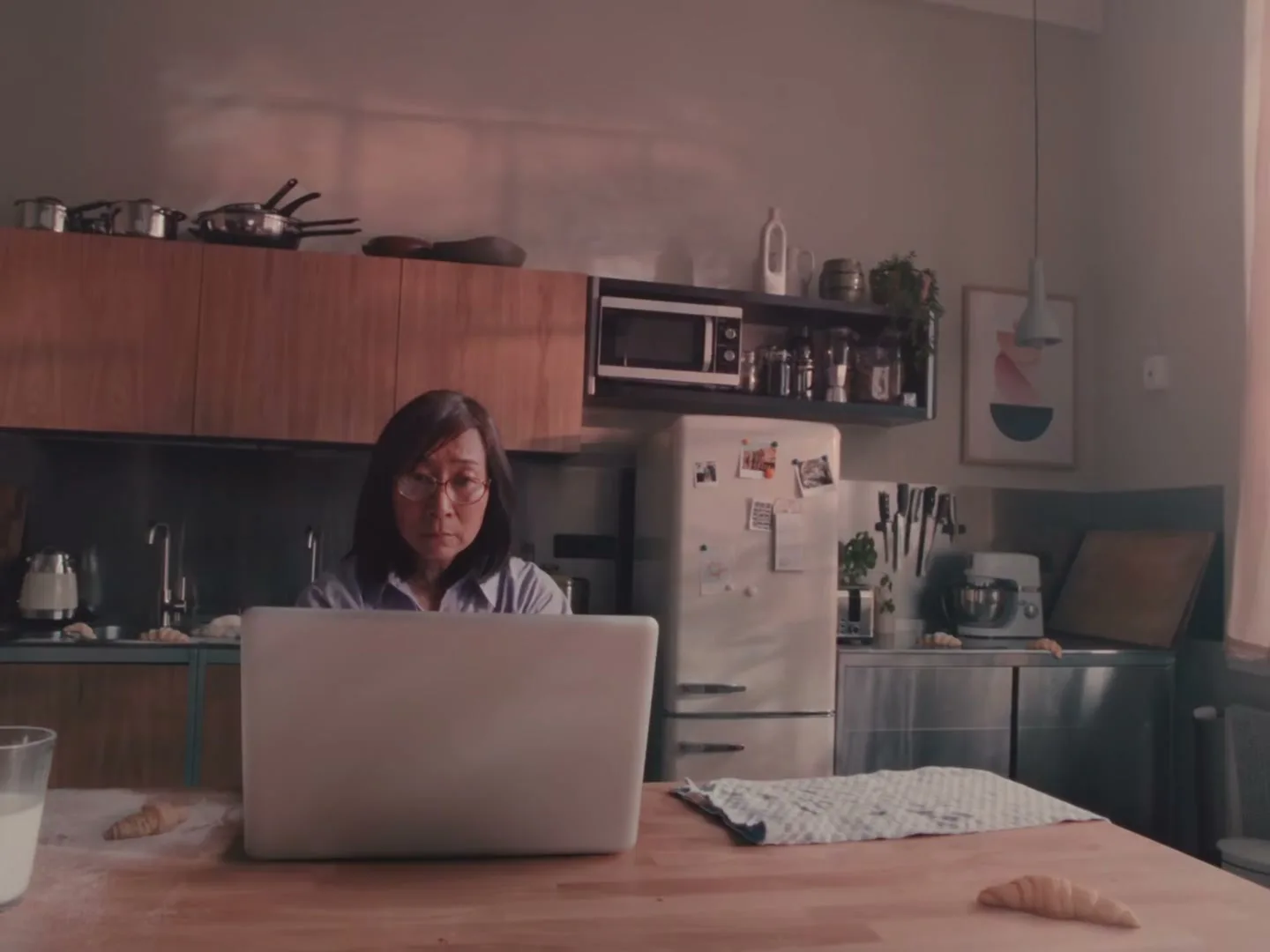 A middle-aged Asian woman with glasses sits at a kitchen counter, looking intently at a laptop screen, with baking ingredients around her.