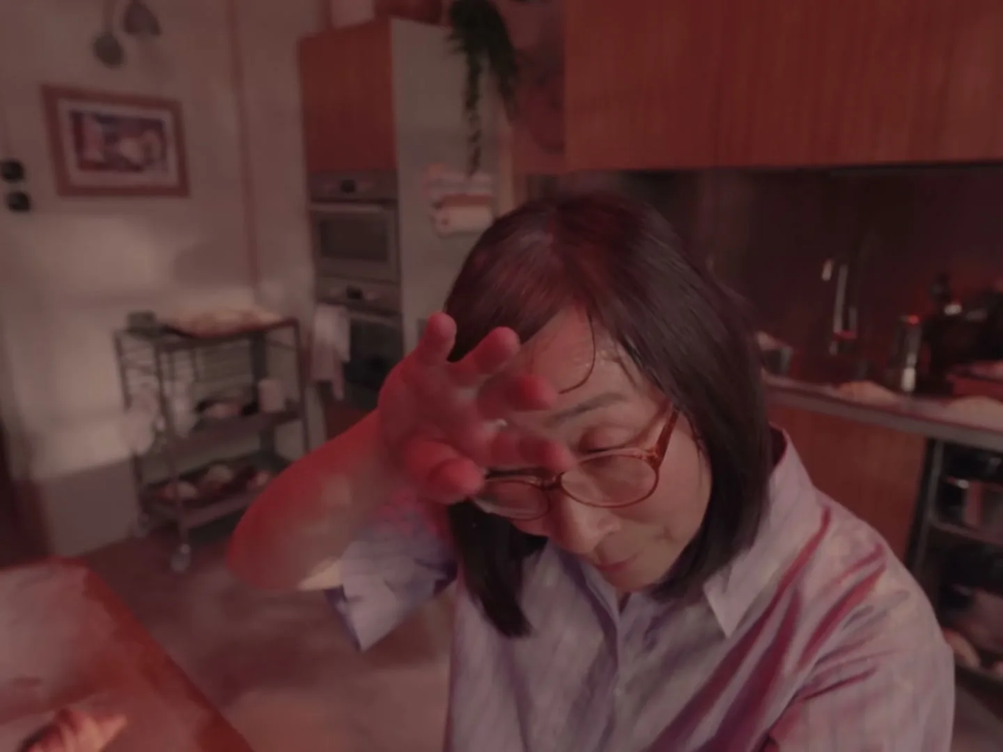 A woman with glasses and a striped shirt wipes her brow in a kitchen, looking tired and stressed under red lighting.