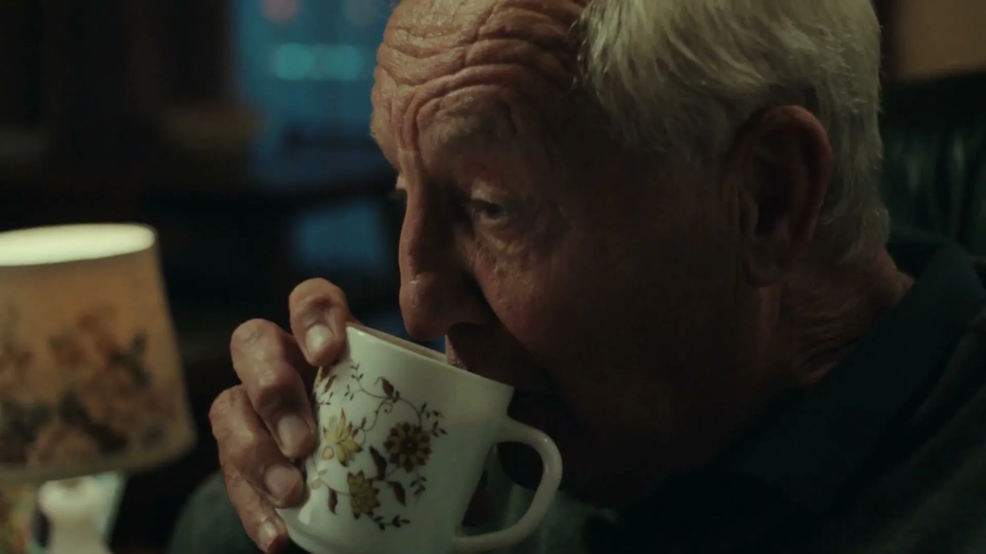 An elderly man with white hair sips from a floral mug in a dimly lit interior, with a lamp visible to his left.