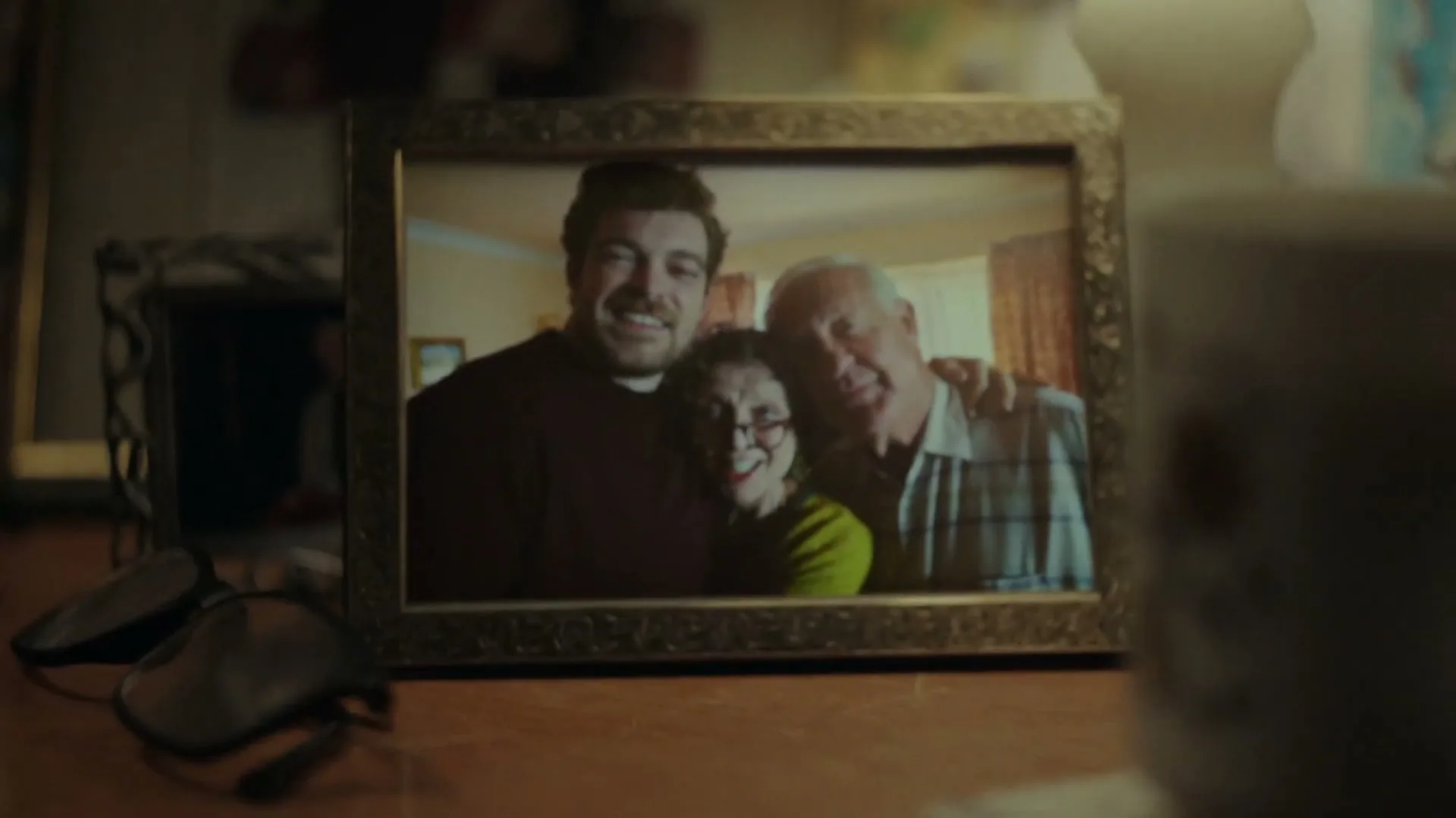 A framed photo of a young man, an older woman, and an older man smiling, is seen on a wooden table in a cozy interior.