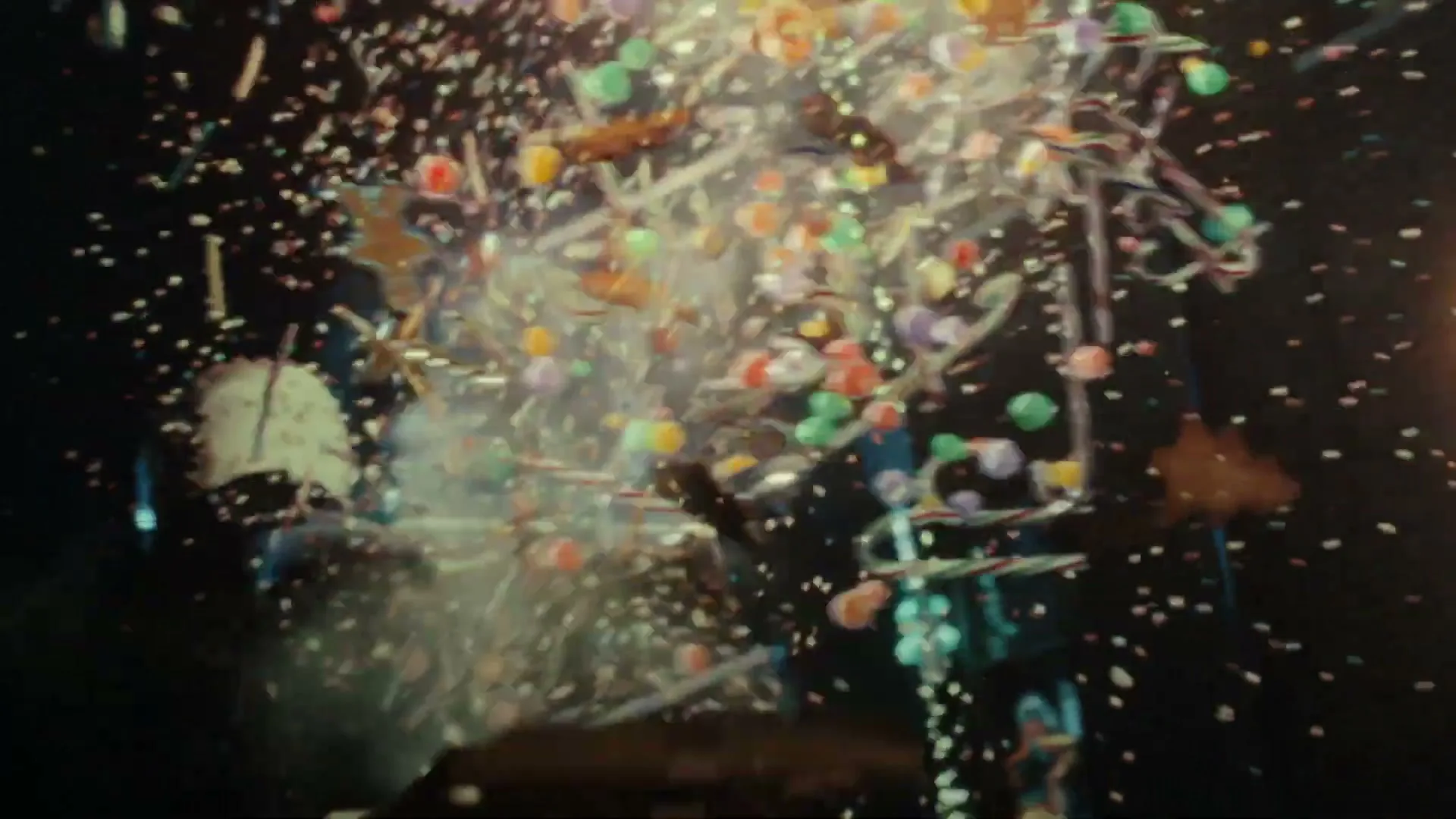 A brown box with white polka dots explodes, sending candy canes, colorful candies, and confetti flying into the air against a dark background.