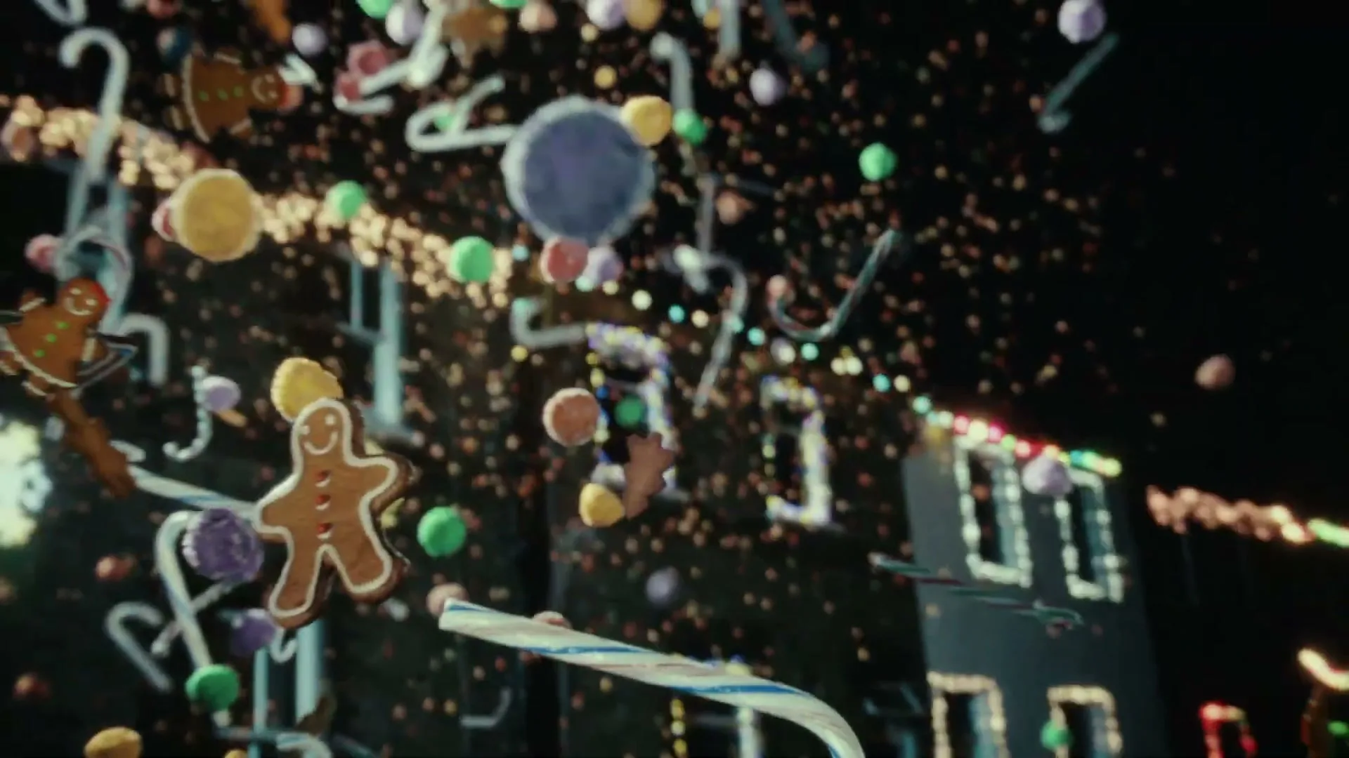 A gingerbread man, candy canes, and colorful candies float in front of a dark building decorated with Christmas lights at night.