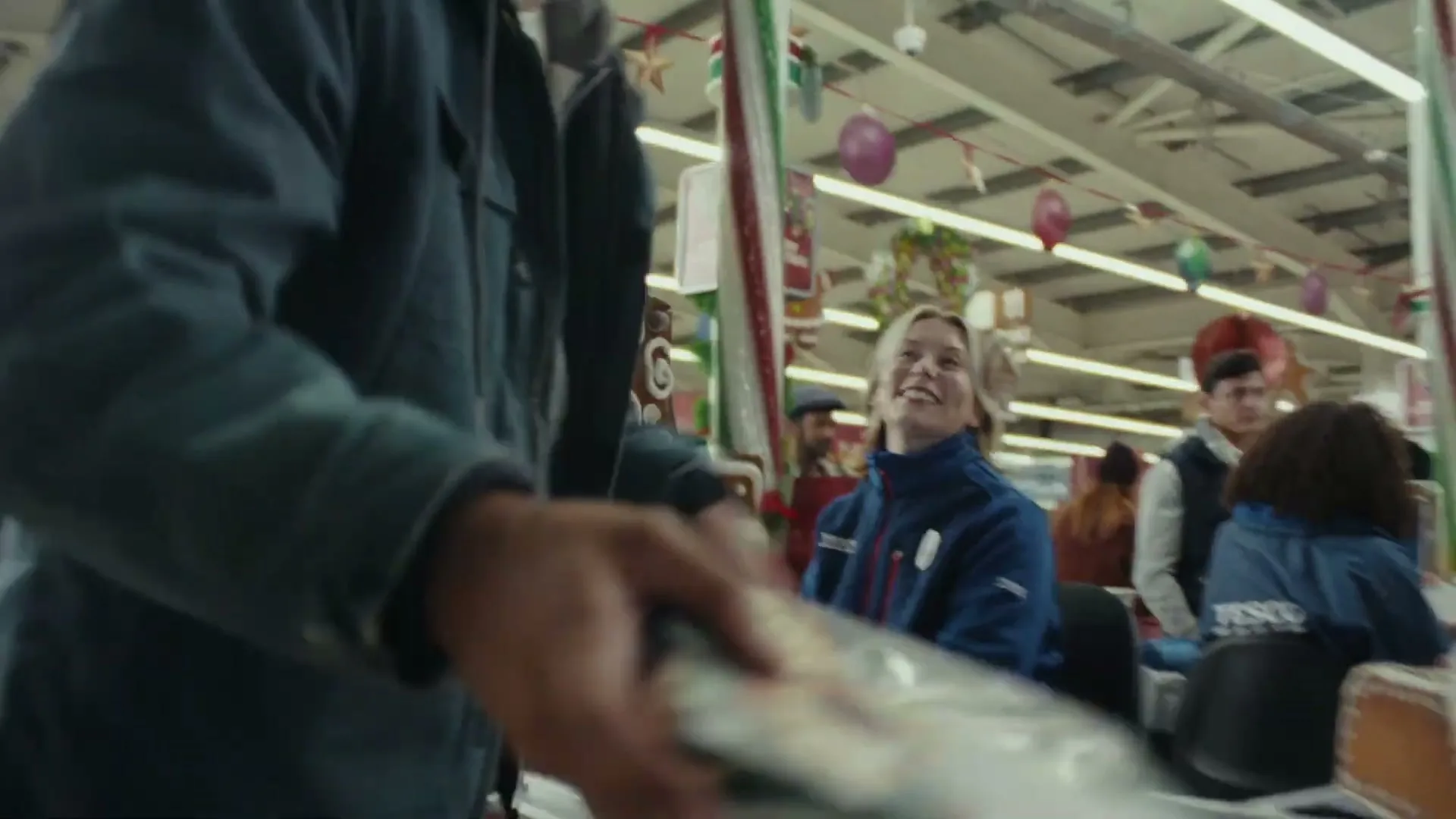 A male customer in a blue jacket places items into a shopping cart at a Tesco checkout, while a female cashier smiles at him.