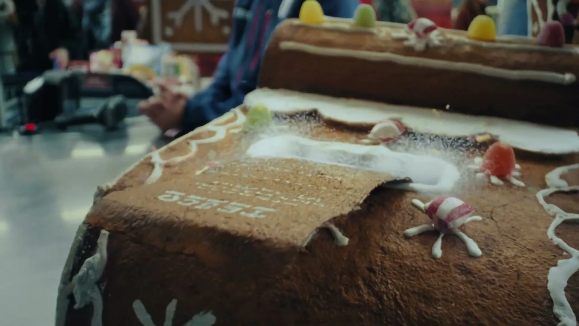 A gingerbread house, decorated with candies and icing, has a gingerbread receipt unrolling from its roof, with a child's hand in the background.