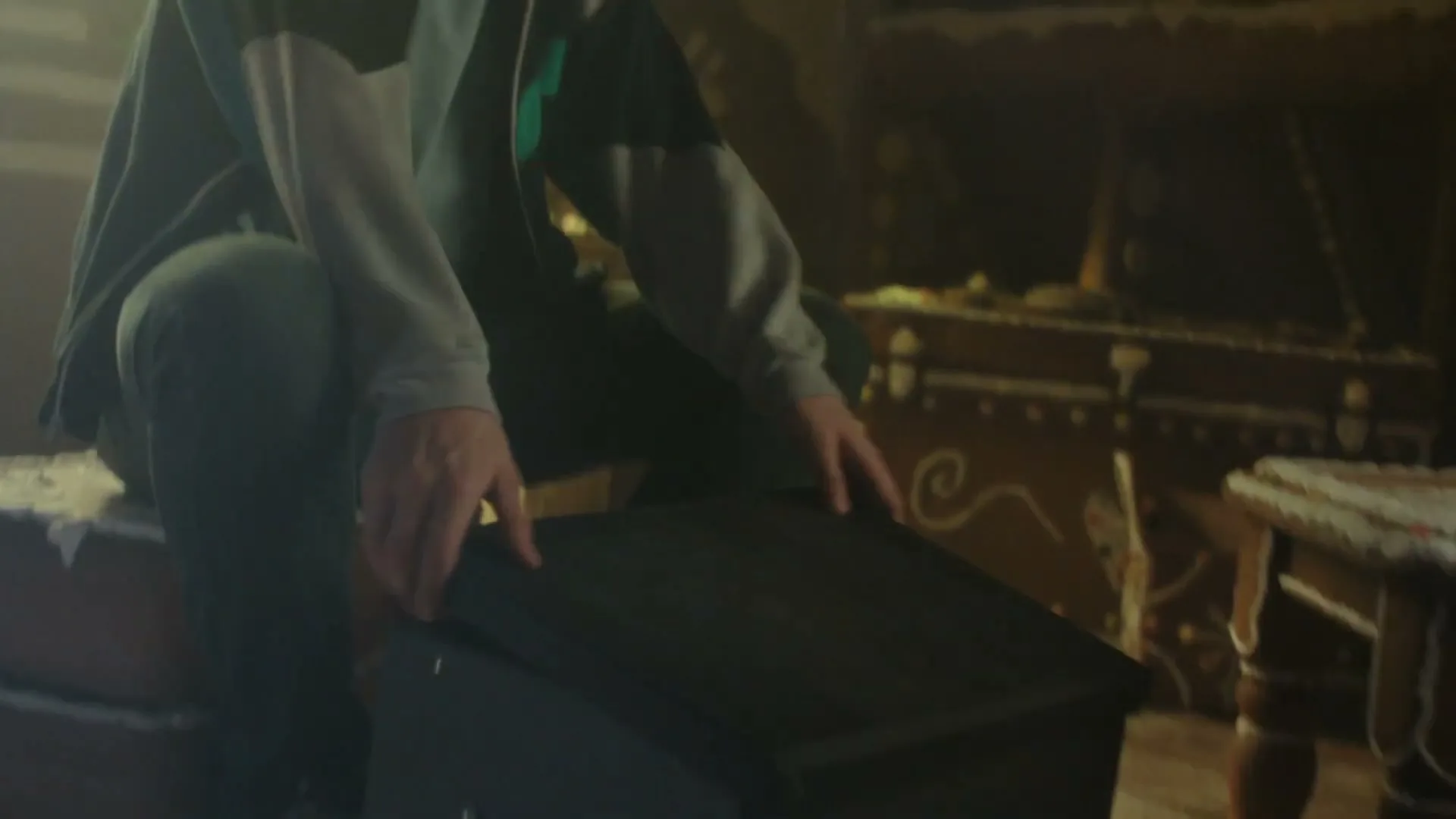 A young man in a hoodie crouches down to open a black box on a wooden floor in a dimly lit room with decorative furniture.