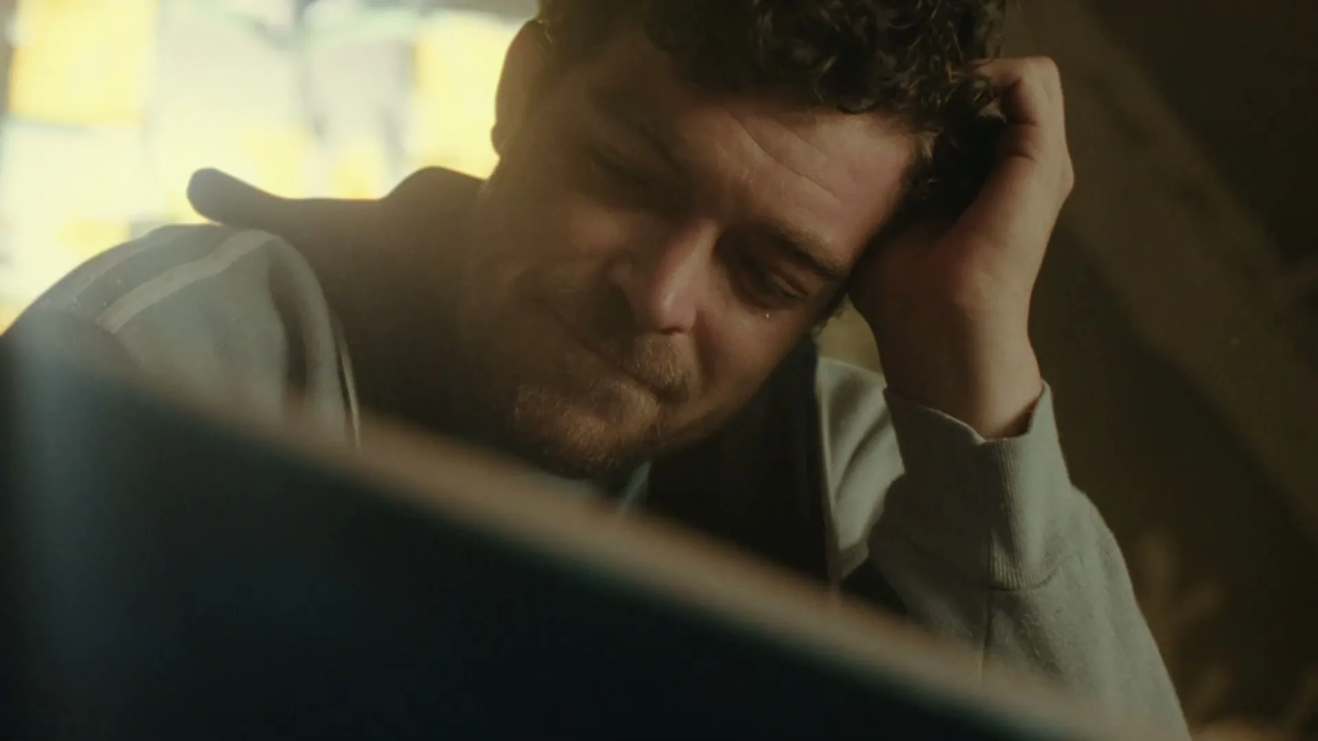 A man with curly hair and a beard, wearing a grey hoodie, rests his head on his hand, looking tired in a dimly lit room.