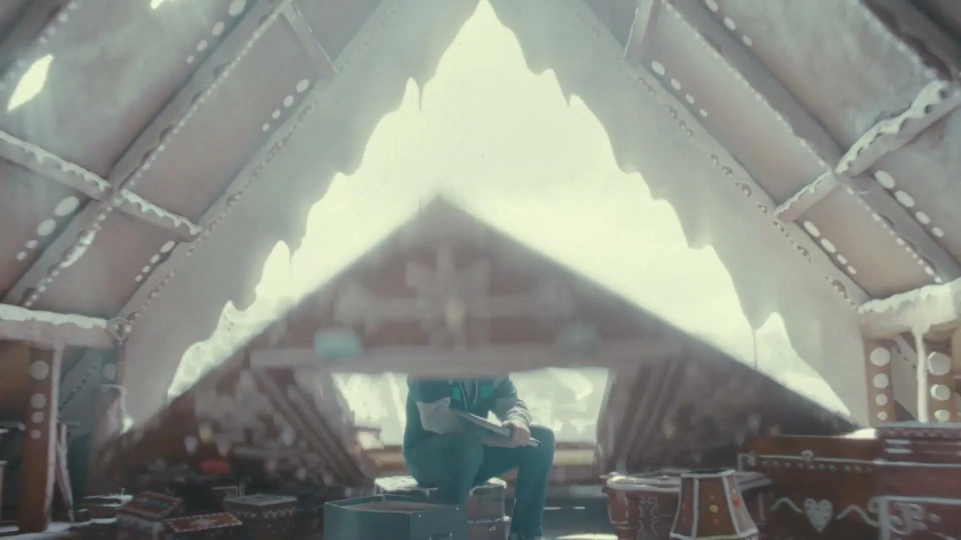 A man in a blue and grey tracksuit sits in a gingerbread house, looking at a photo album, as the room transforms into an icy attic.