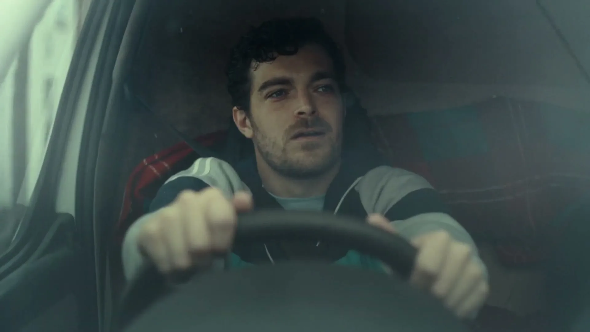 A man with dark curly hair and a beard sits in a car, looking out the windshield with a concerned expression.