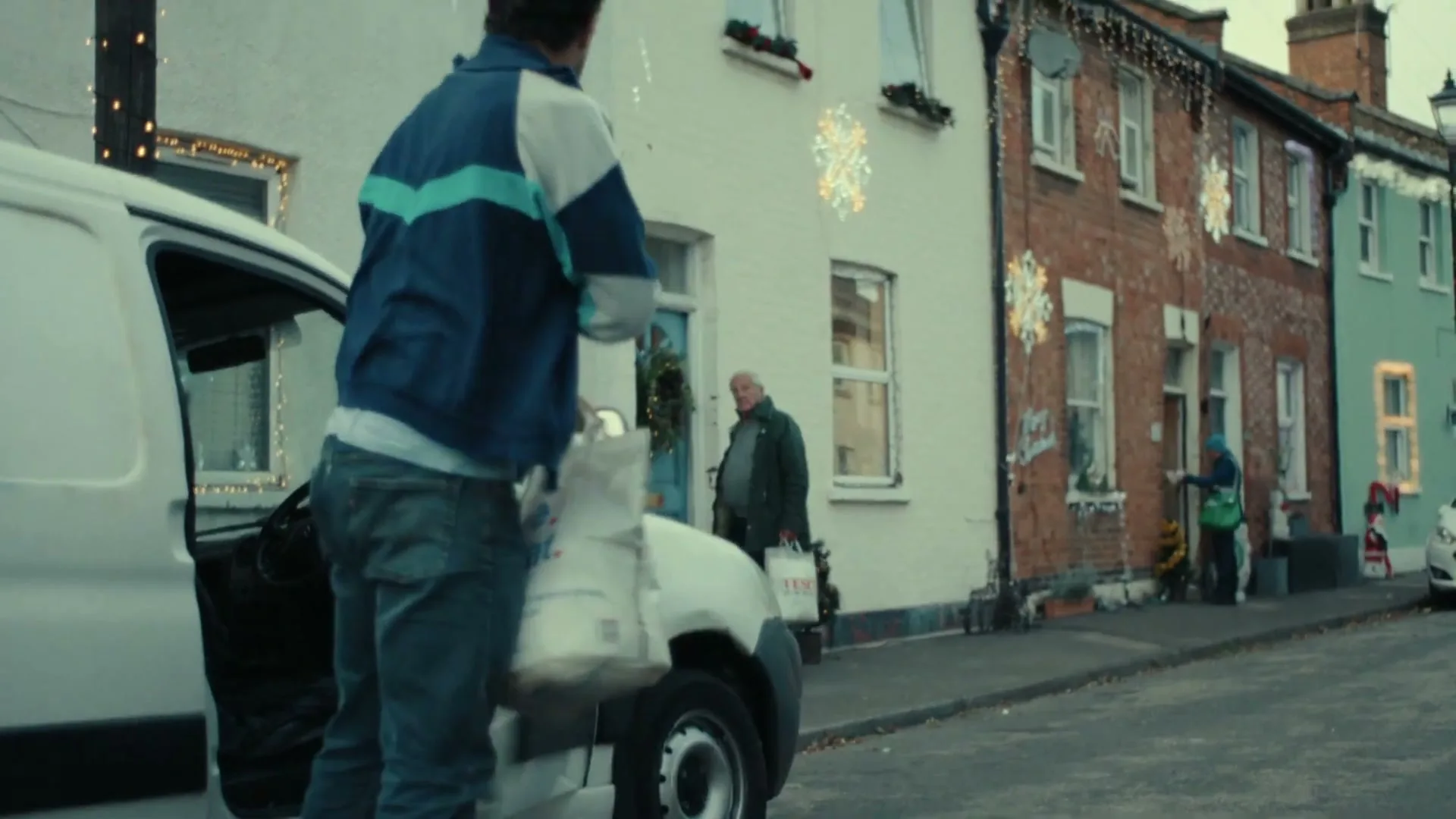 A delivery driver in a blue and white jacket gets out of a white van, holding a white grocery bag, on a street decorated for Christmas.