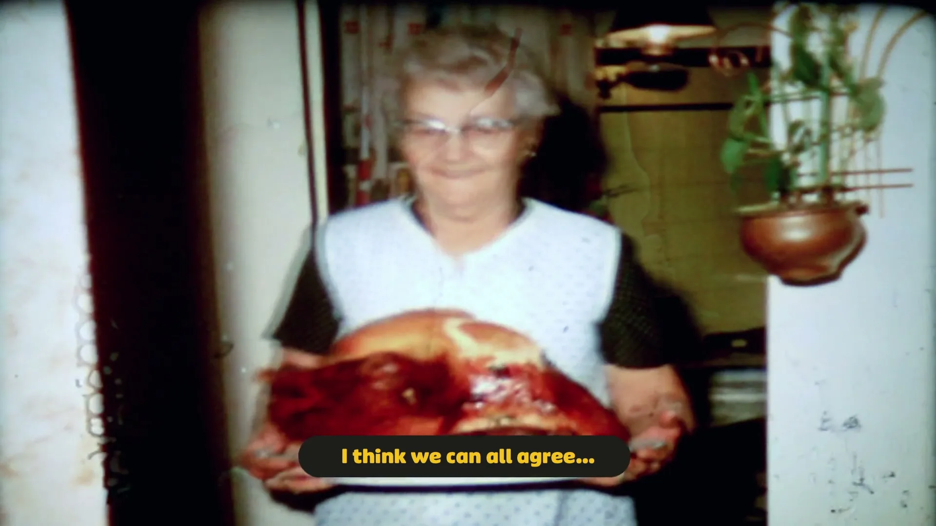 A smiling older woman in glasses and a white apron holds a large roasted turkey on a platter in a home kitchen.