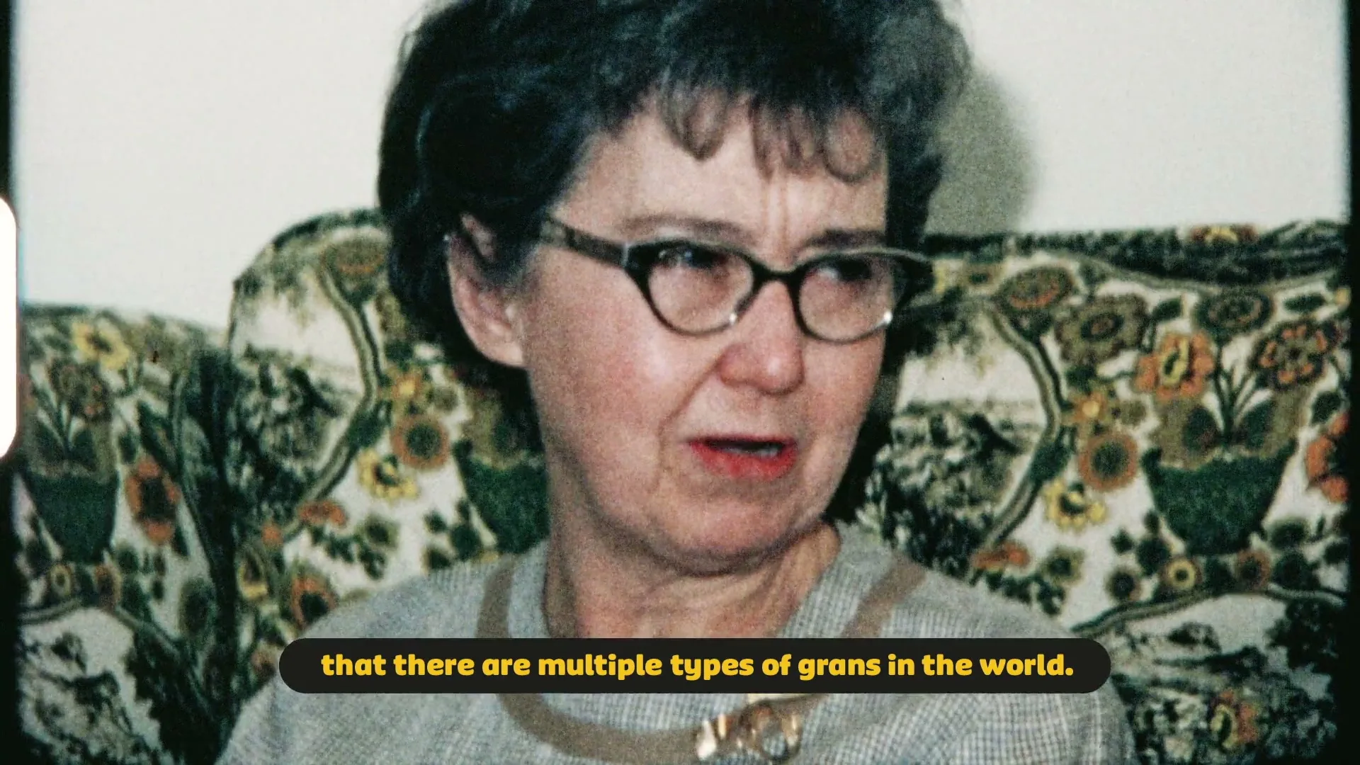 A senior woman with glasses and short grey hair sits on a floral couch, speaking with a slight smile in a medium closeup shot.