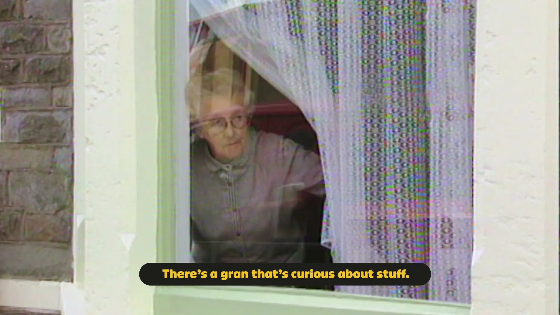 An elderly woman with glasses and a grey top peeks out from behind a lace curtain in a window, looking curious.