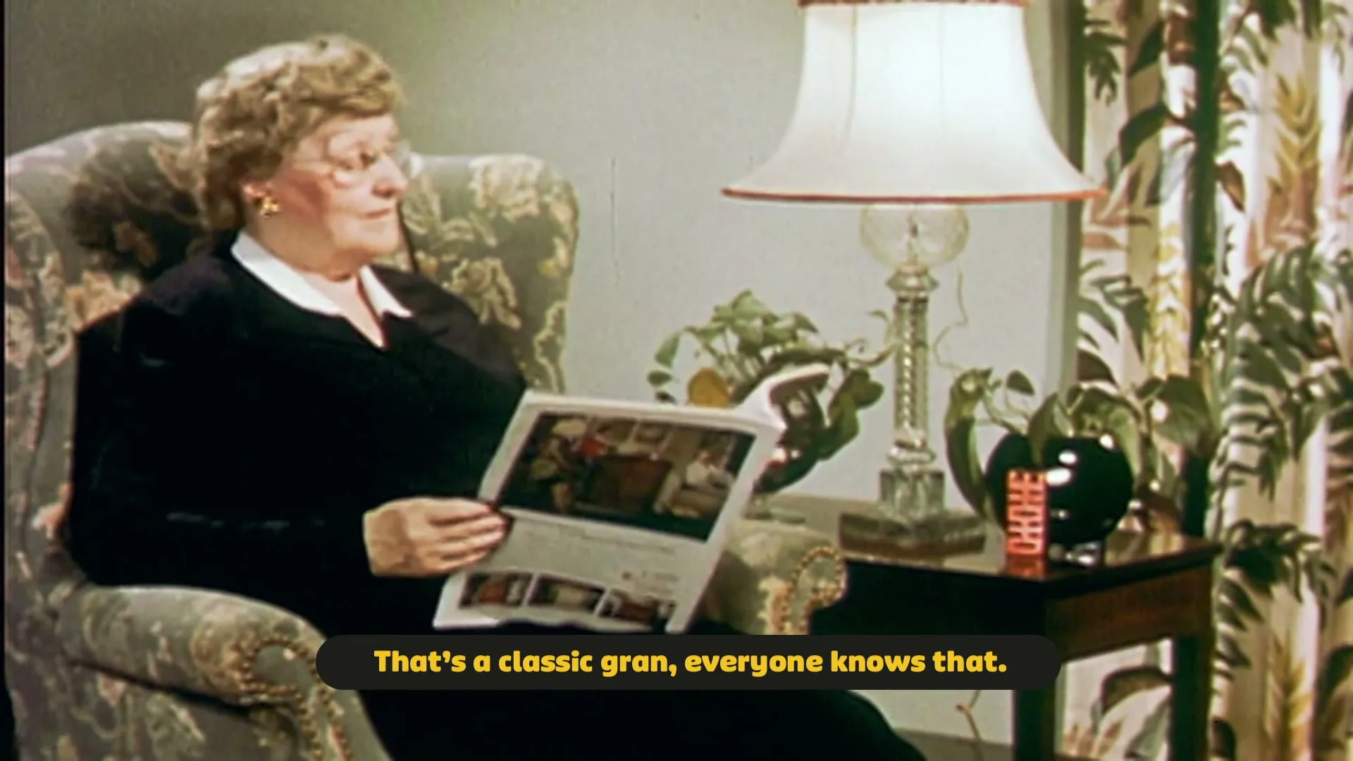 An older woman with curly hair and glasses sits in an armchair, reading a magazine in a cozy living room.
