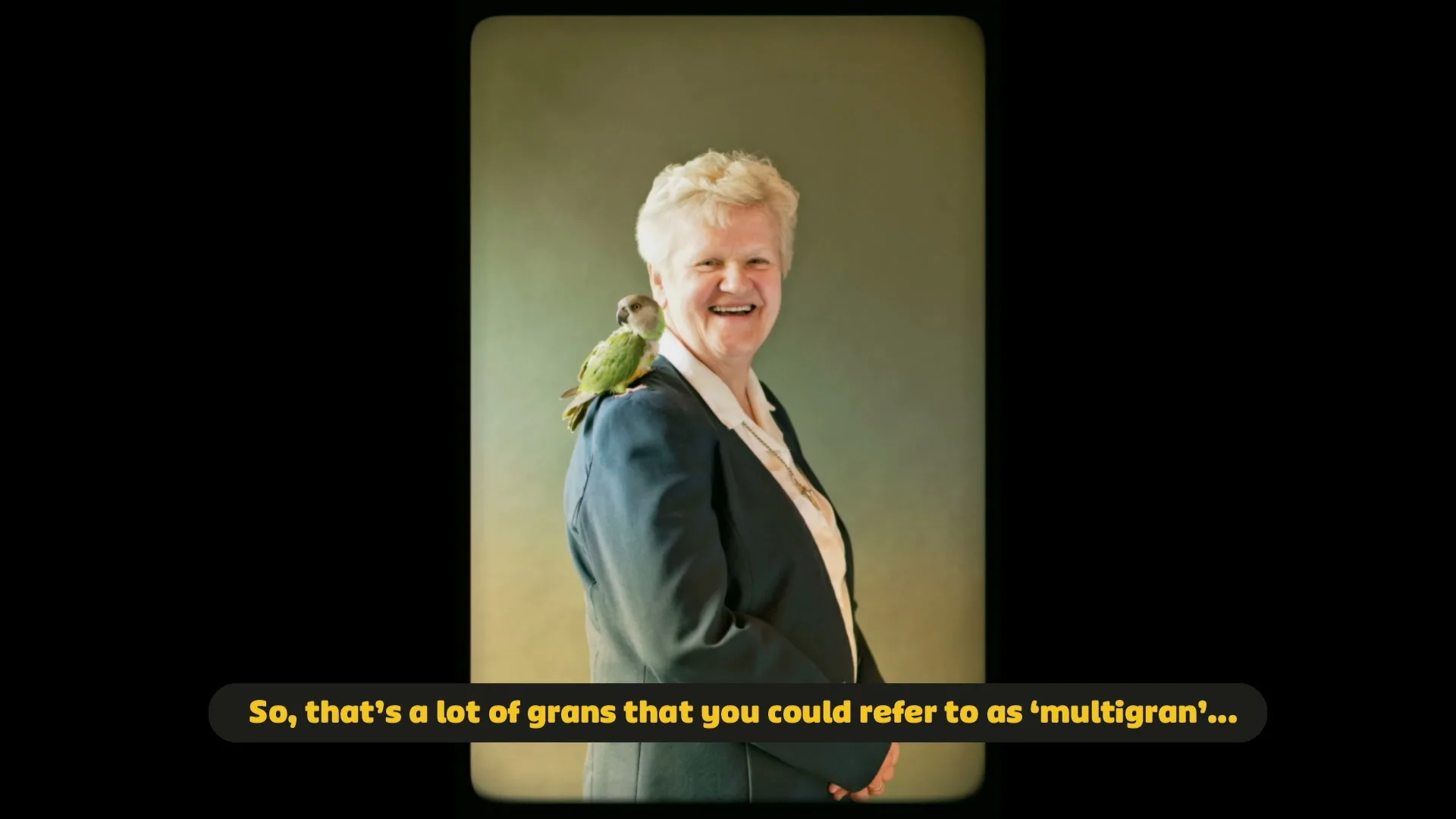 A smiling older woman with short blonde hair and a green parrot on her shoulder stands against a green background, looking at the camera.