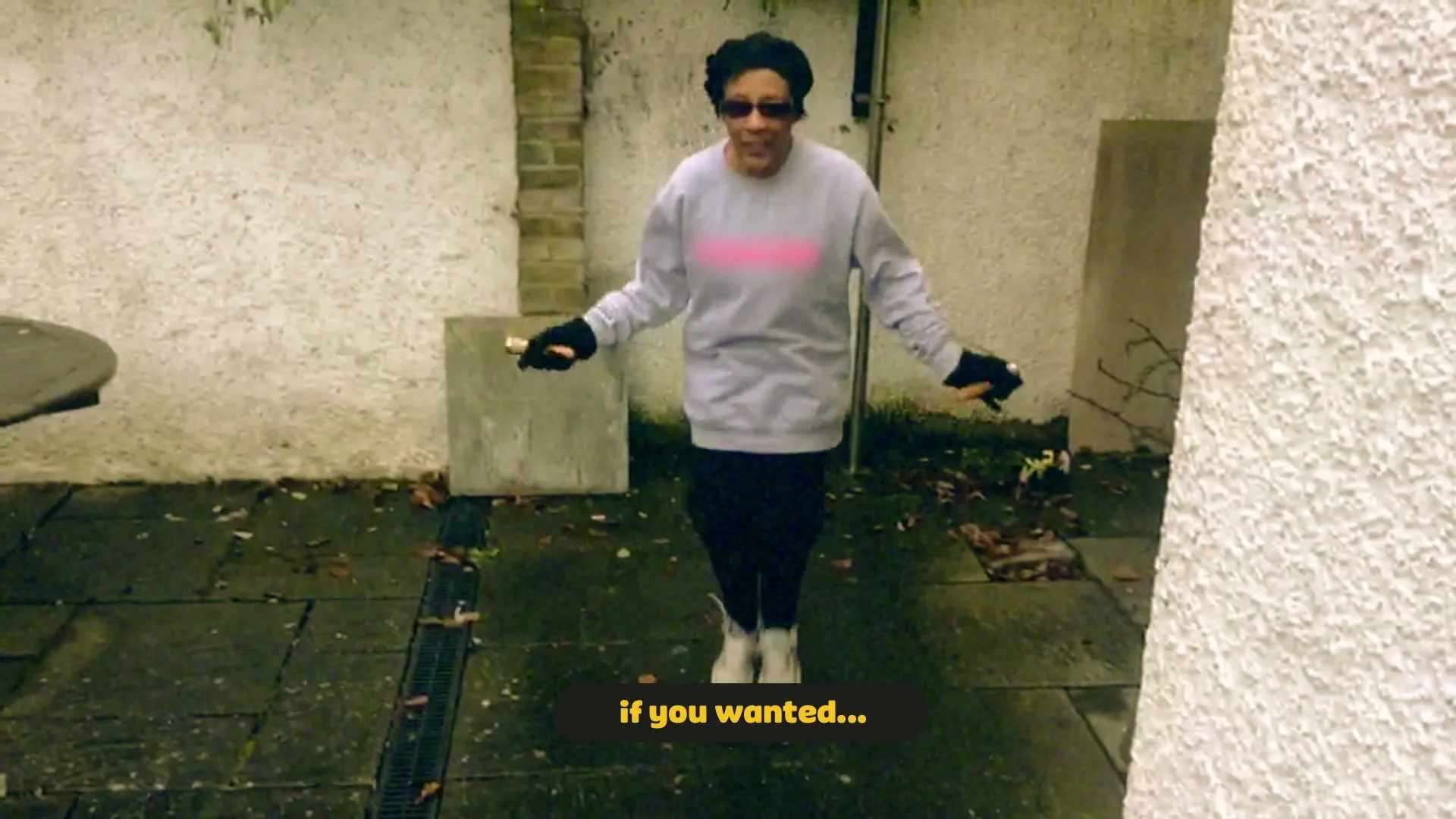 A woman in a grey sweatshirt and black pants jumps rope in a paved backyard with white walls, smiling and wearing sunglasses.