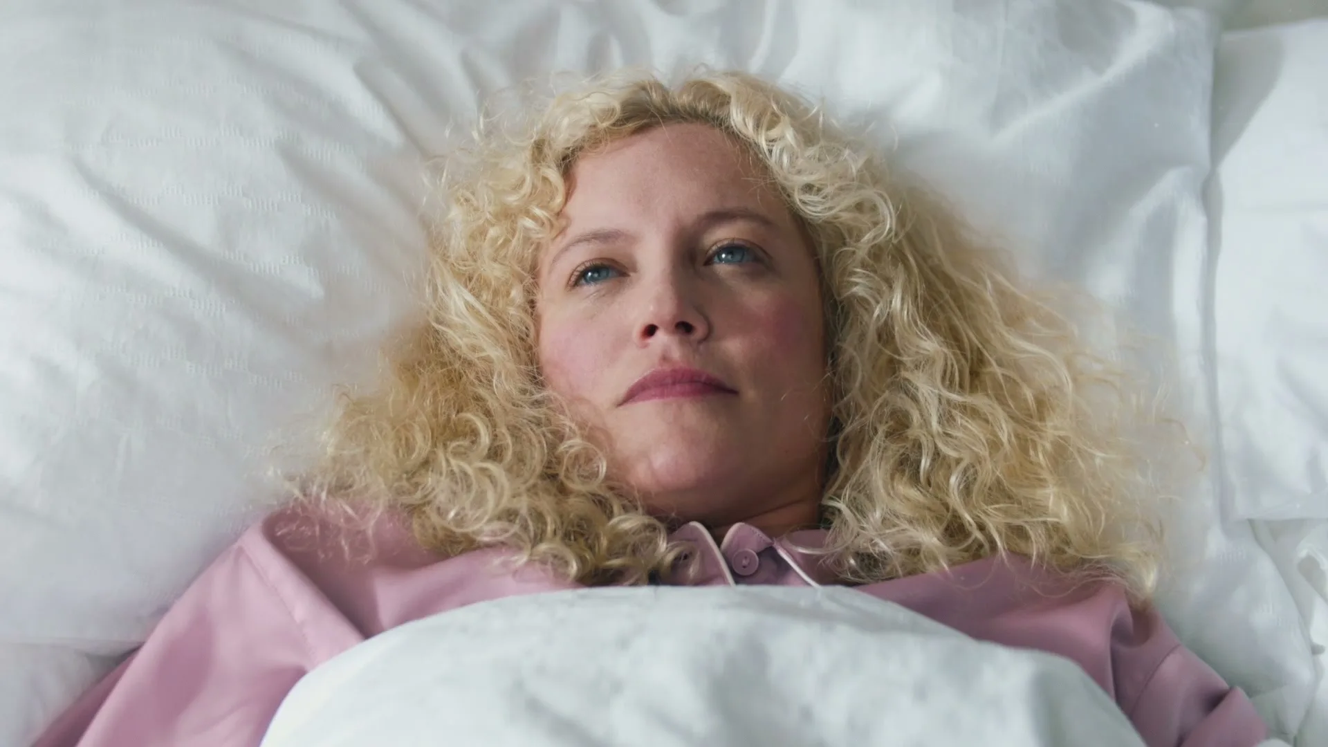 A young woman with curly blonde hair lies in bed, looking up thoughtfully, wearing pink pajamas in a bright bedroom.
