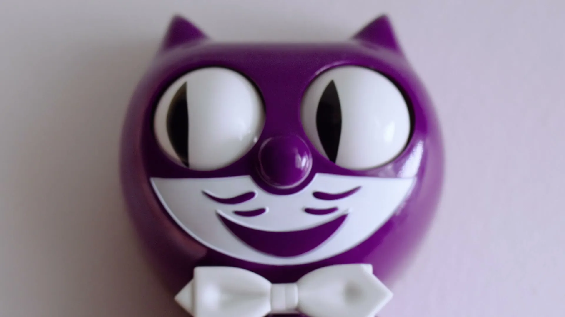 A purple cat-shaped toy with large white eyes and a white bow tie is shown in a close-up shot against a light background.