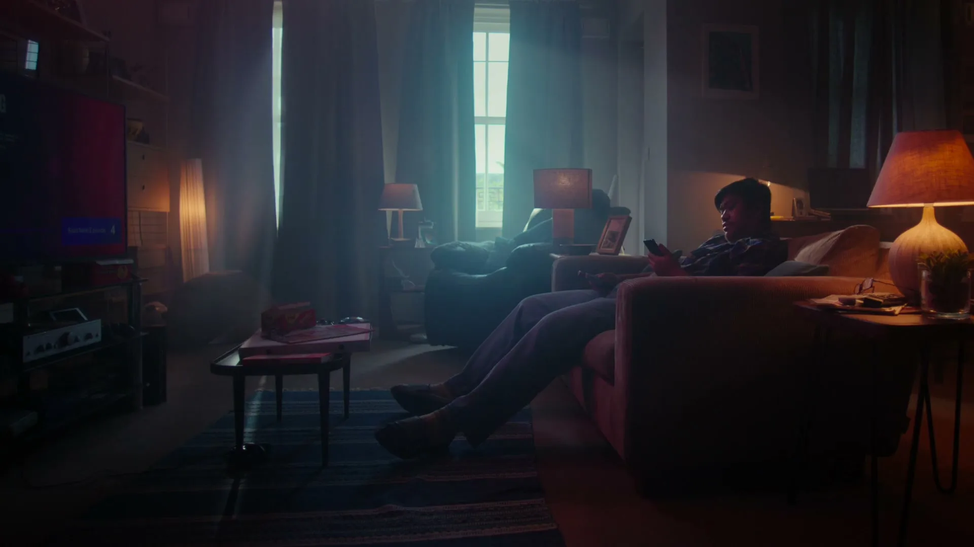 A young man in a plaid shirt relaxes on a sofa, looking at his phone in a dimly lit living room.