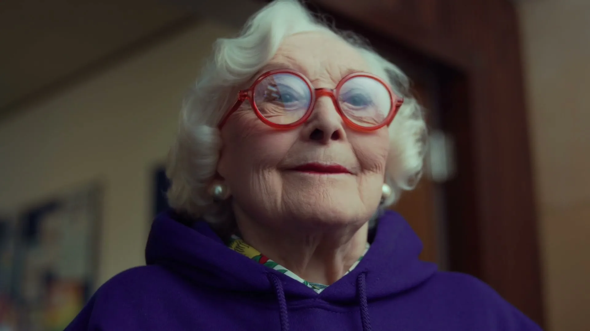 An elderly woman with white hair and red glasses smiles slightly, looking up and to the right in an interior setting.