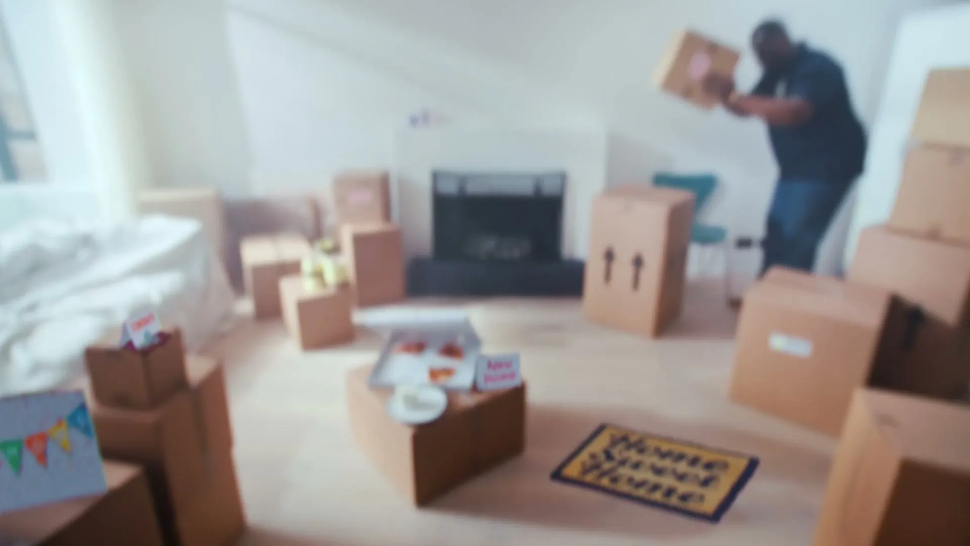 A man in a dark t-shirt pushes a moving box across a bright, empty living room with a fireplace and many cardboard boxes.