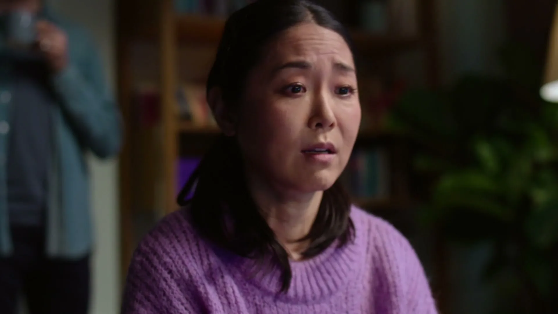 A young Asian woman in a purple sweater looks distressed while a man stands blurred in the background of a cozy living room.