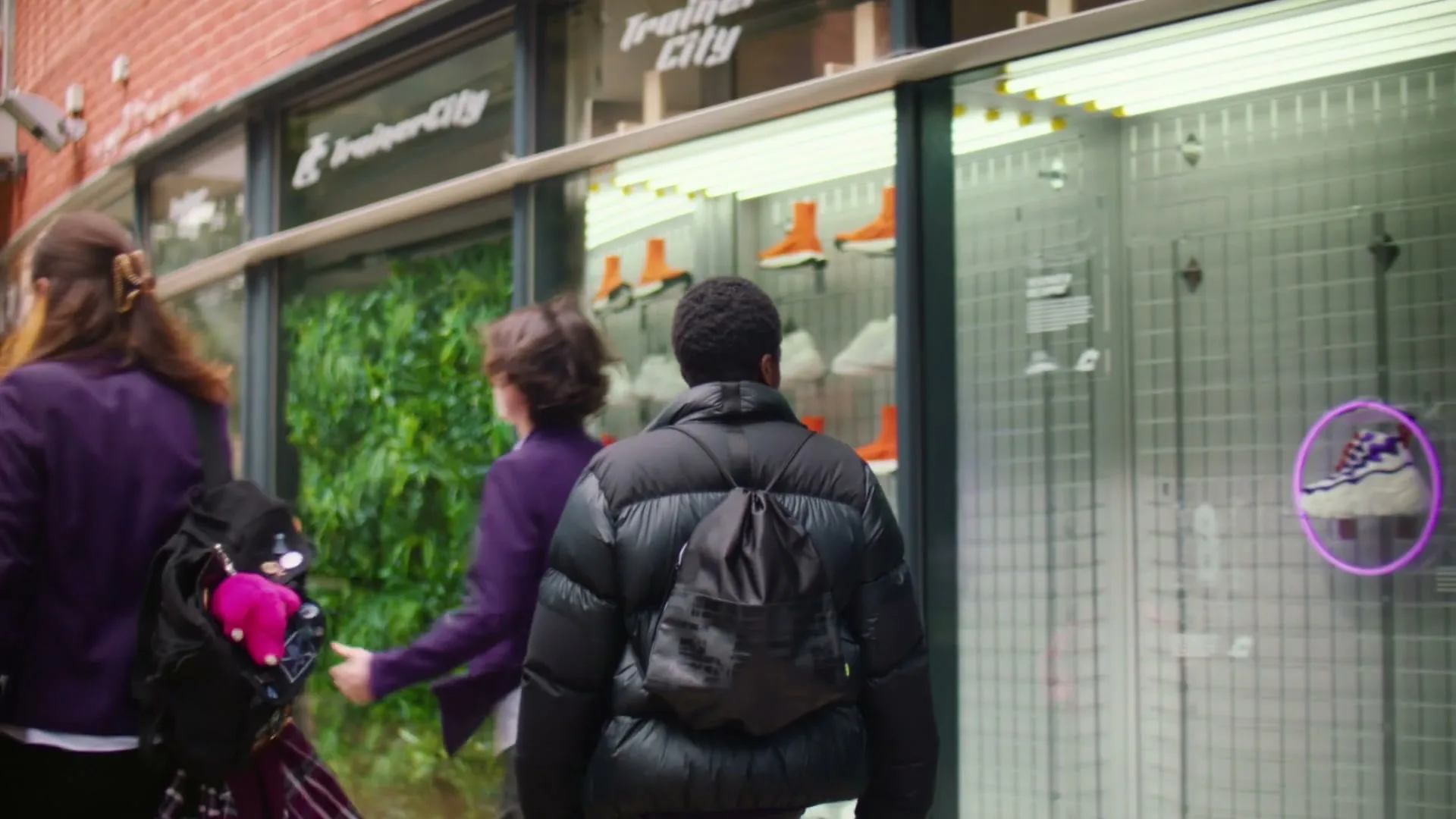 Three teens walk past a shoe store window, one wearing a puffer jacket and backpack, on a city street.