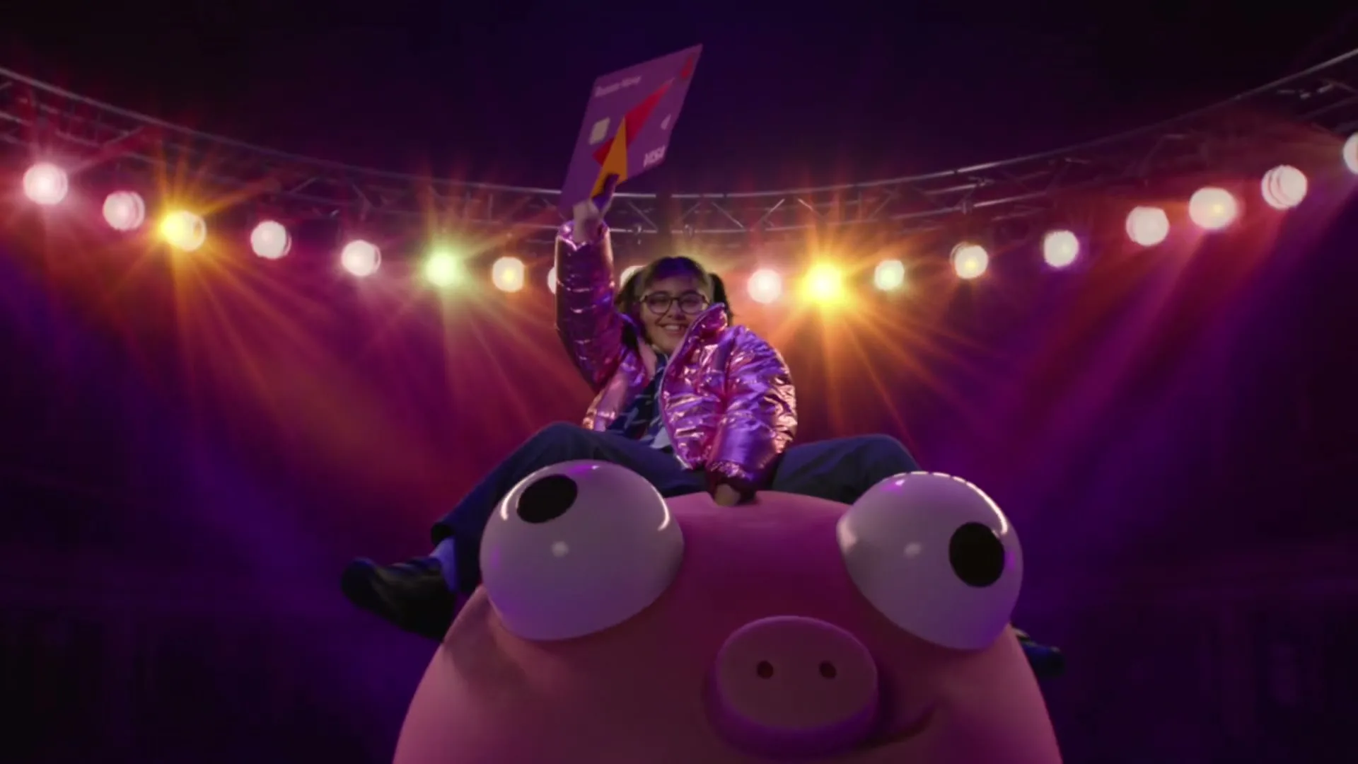 A young girl in a shiny pink jacket sits atop a giant pink piggy bank, holding up a purple card, under bright stage lights.