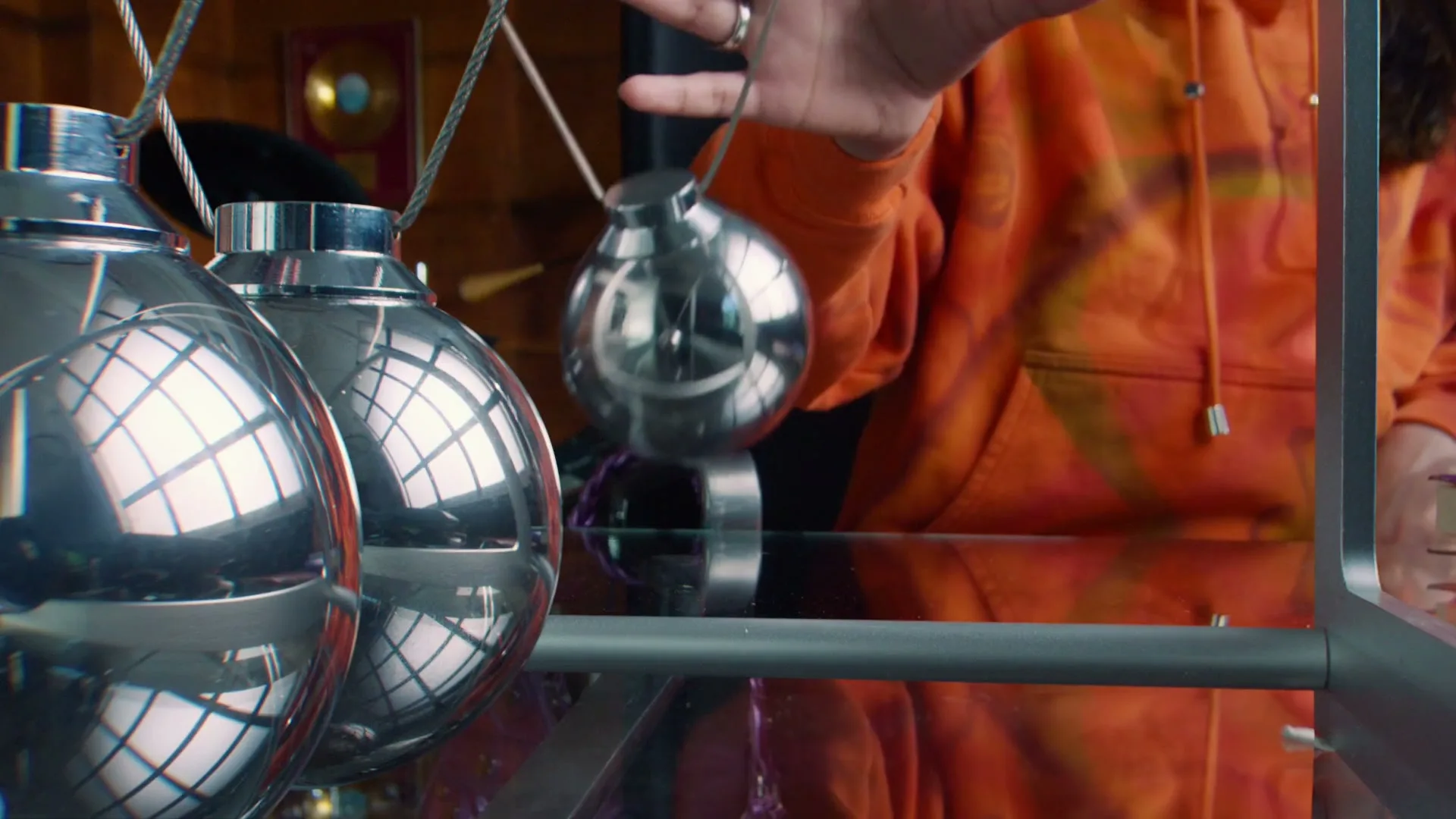 A person in an orange hoodie holds a chrome sphere, part of a Newton's Cradle, in a room with warm lighting.