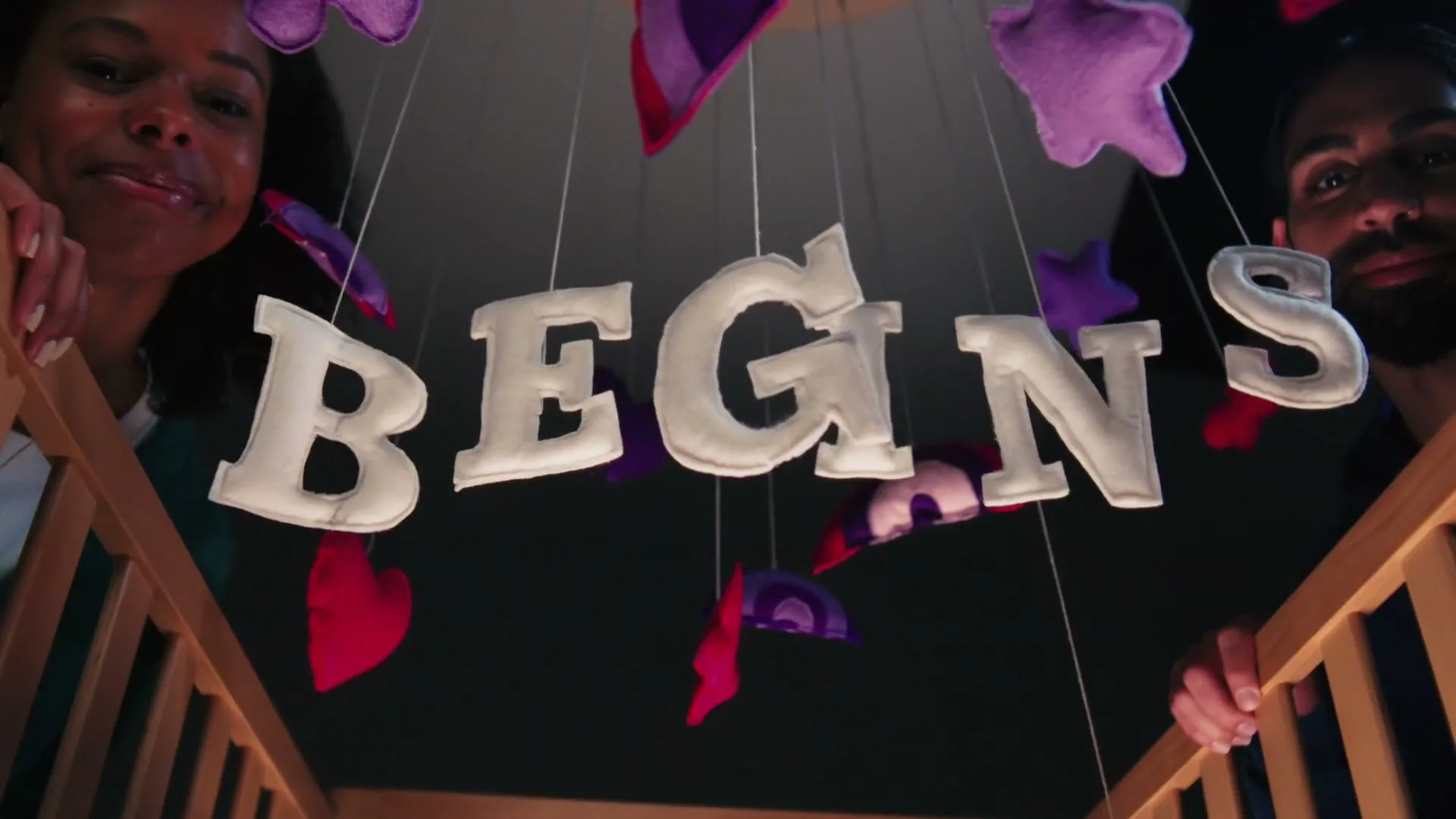 A couple looks down into a baby's crib, where a mobile spells out "BEGINS" in felt letters, surrounded by colorful shapes.
