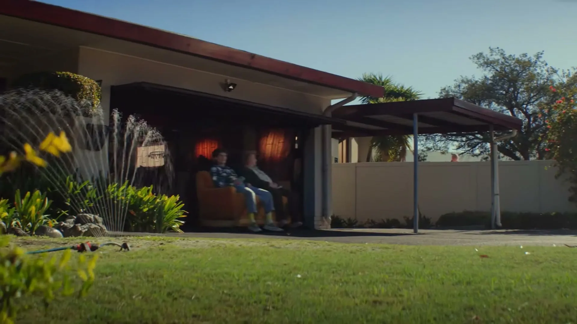 Two young men sit on a couch that slides out of a garage, past a sprinkler, onto a suburban driveway on a sunny day.