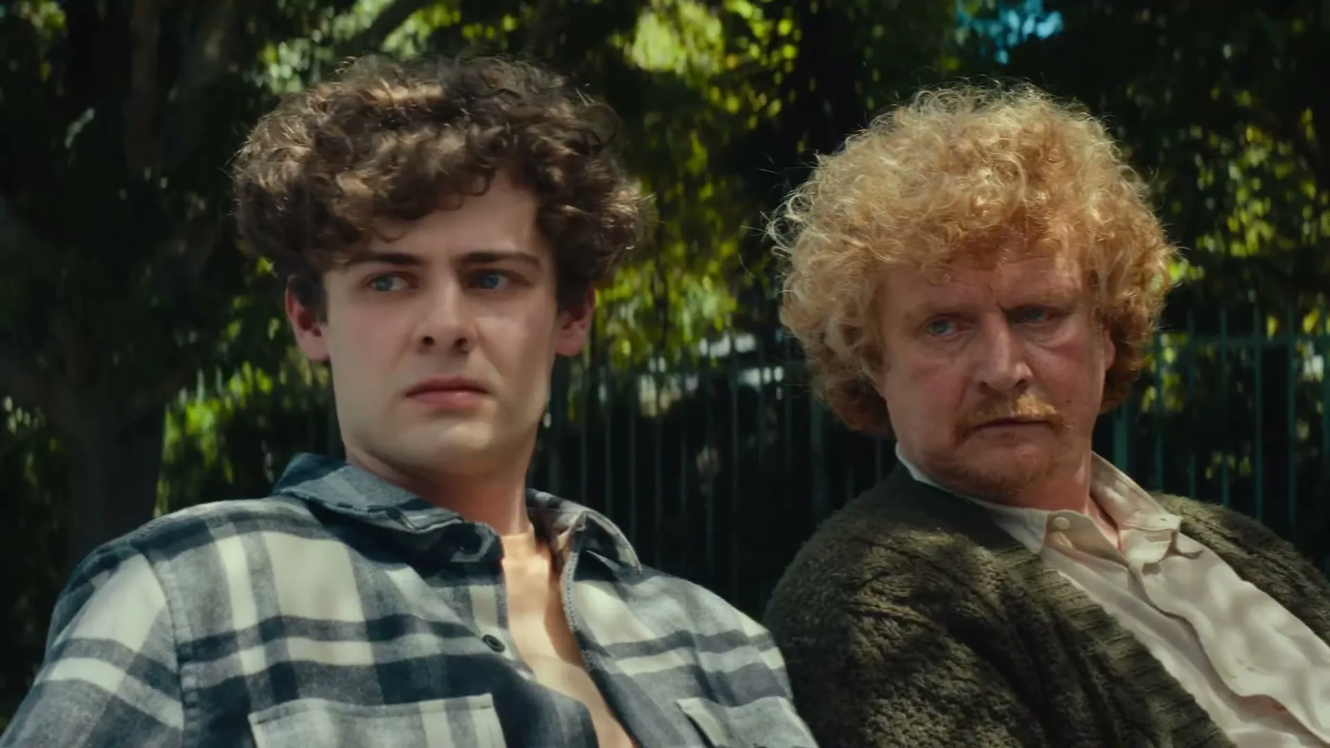 Two men, one young with curly brown hair and one older with curly blonde hair, sit side-by-side, looking forward with serious expressions outdoors.