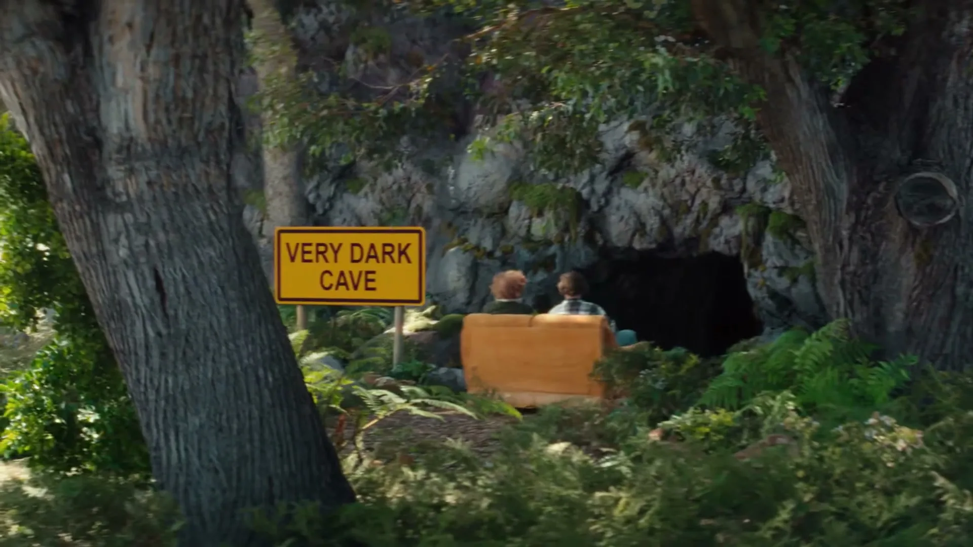 Two young men in a brown cart approach a "Very Dark Cave" sign and enter a cave in a lush, green forest.