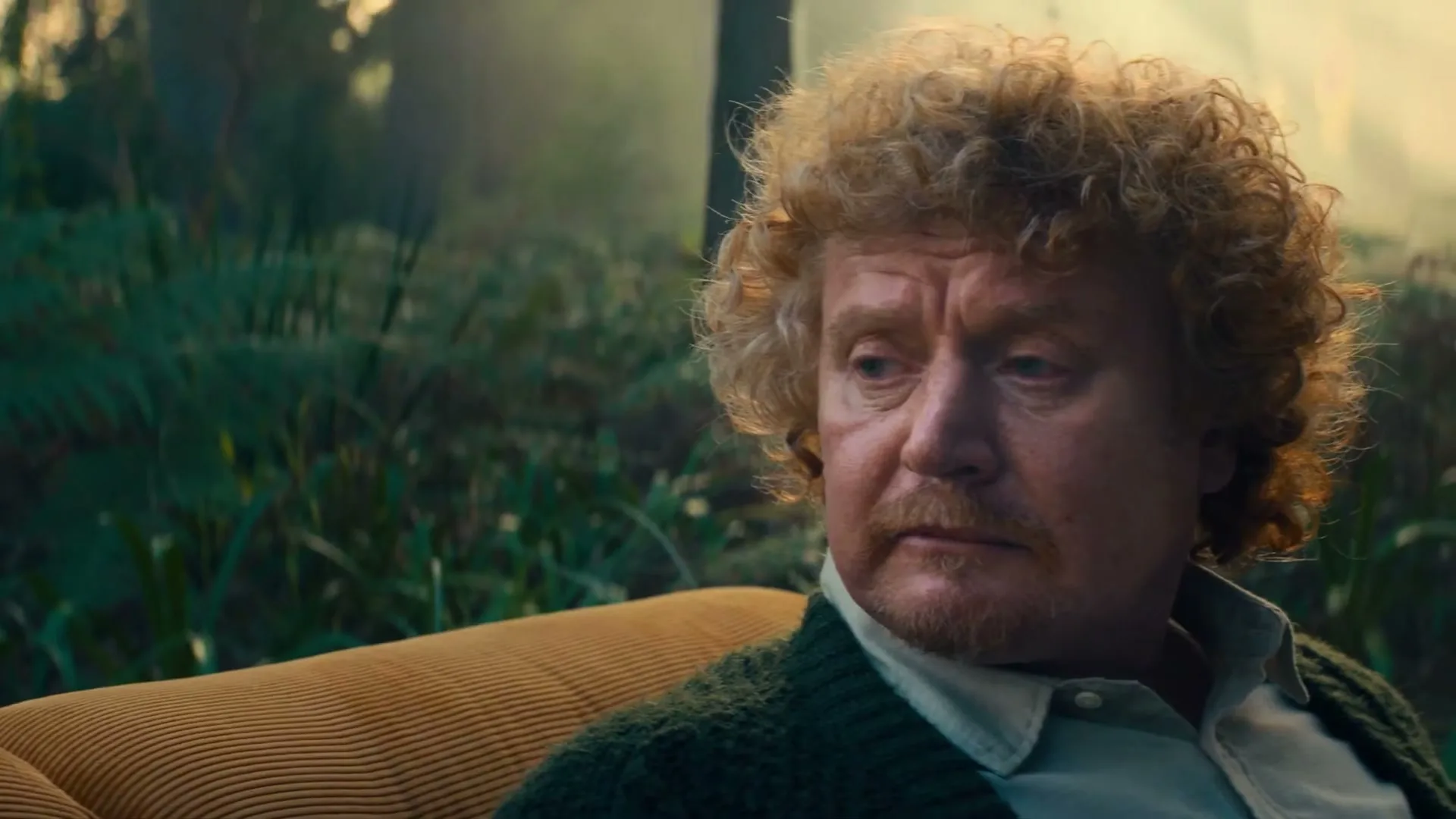 A middle-aged man with curly red hair and a mustache sits on a yellow couch, looking off-screen with a concerned expression in a lush, green, jungle-like setting.