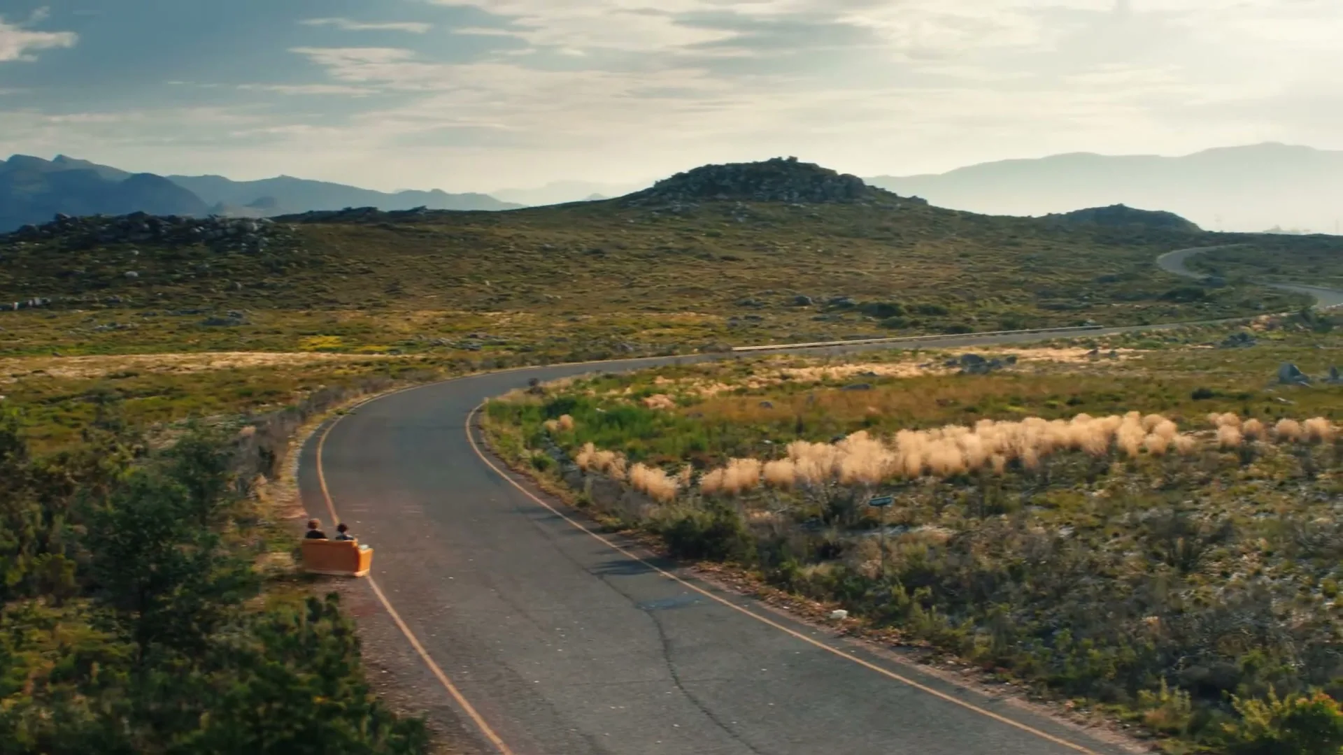 Two people in a small, orange, open-top vehicle drive along a winding road through a vast, grassy landscape with mountains in the distance.
