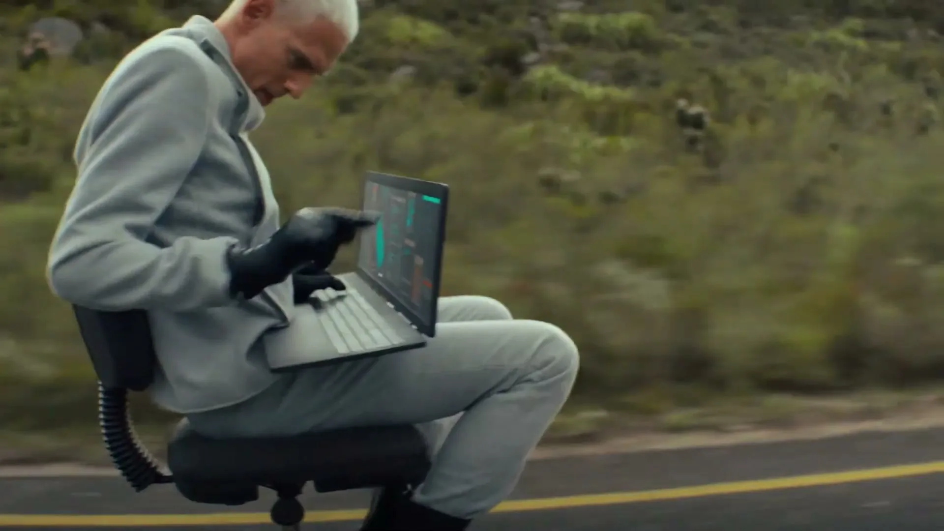 A man with short white hair, wearing a light grey suit and black gloves, sits on an office chair, using a laptop on a road with green hills in the background.