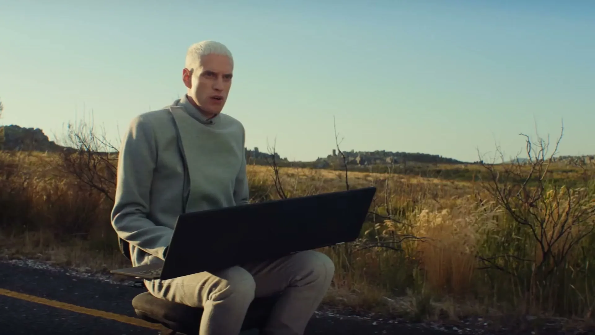 A man with white hair sits on a road in a field, looking at a laptop that he then closes, appearing frustrated.