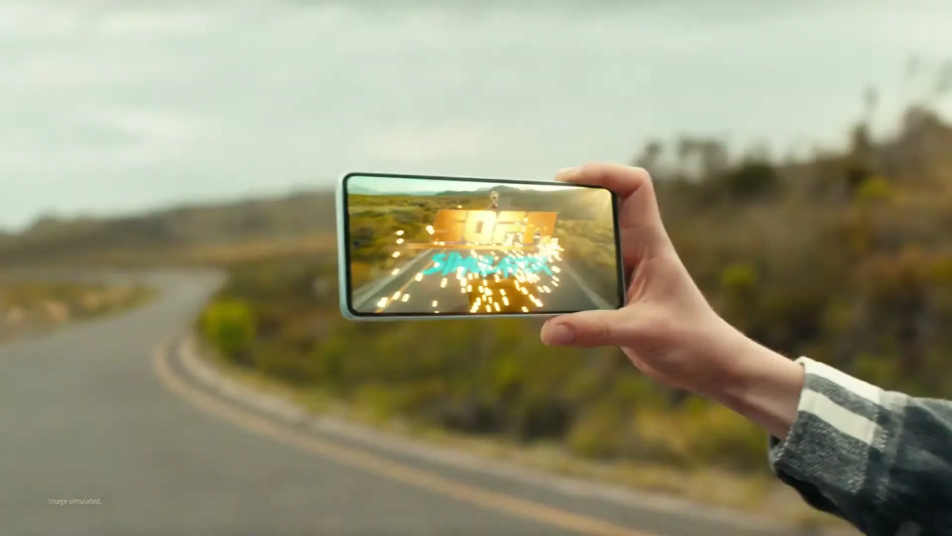 A young man with curly hair smiles while holding a light blue smartphone horizontally, displaying a game with "SOFA SIMULATOR" text on its screen, against a blurred outdoor road background.