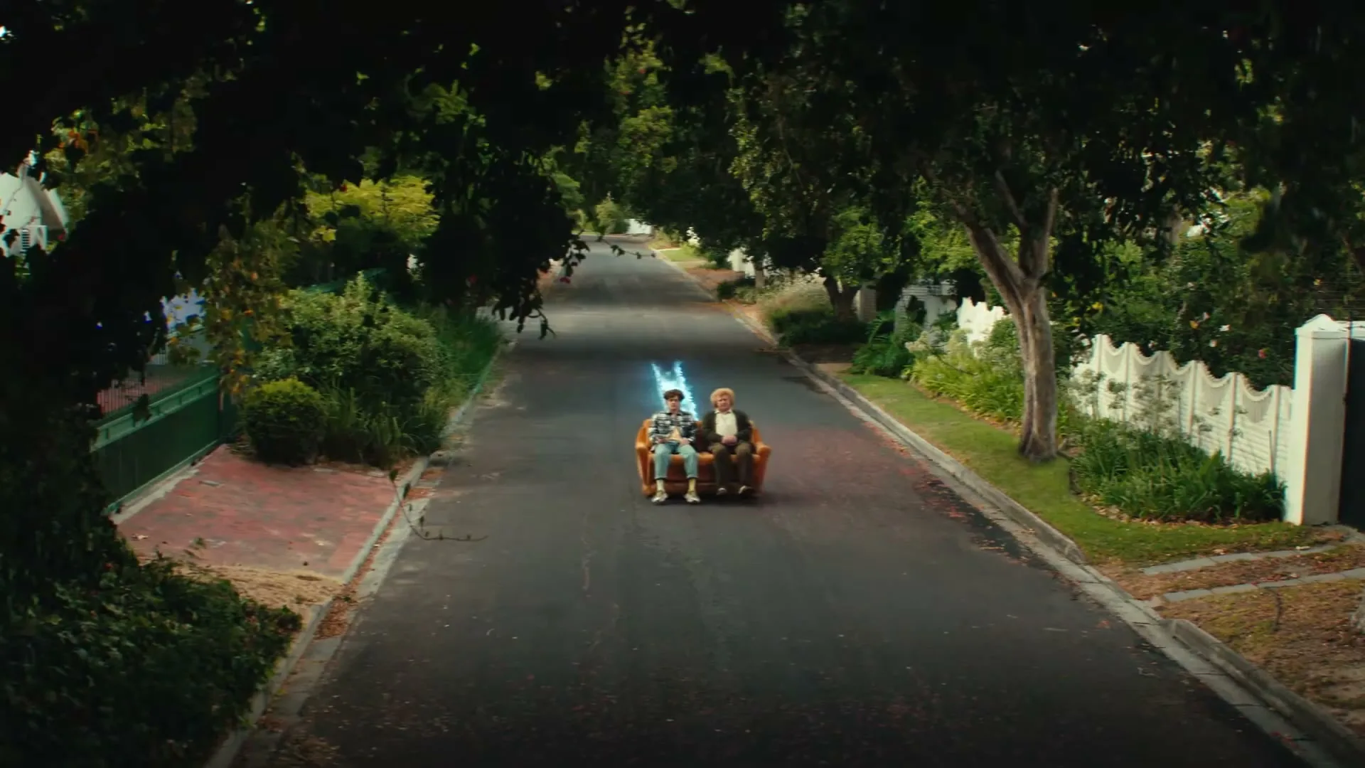 Two men, one young and one older, sit on a couch in the middle of a tree-lined suburban street as a blue energy beam shoots from the couch.