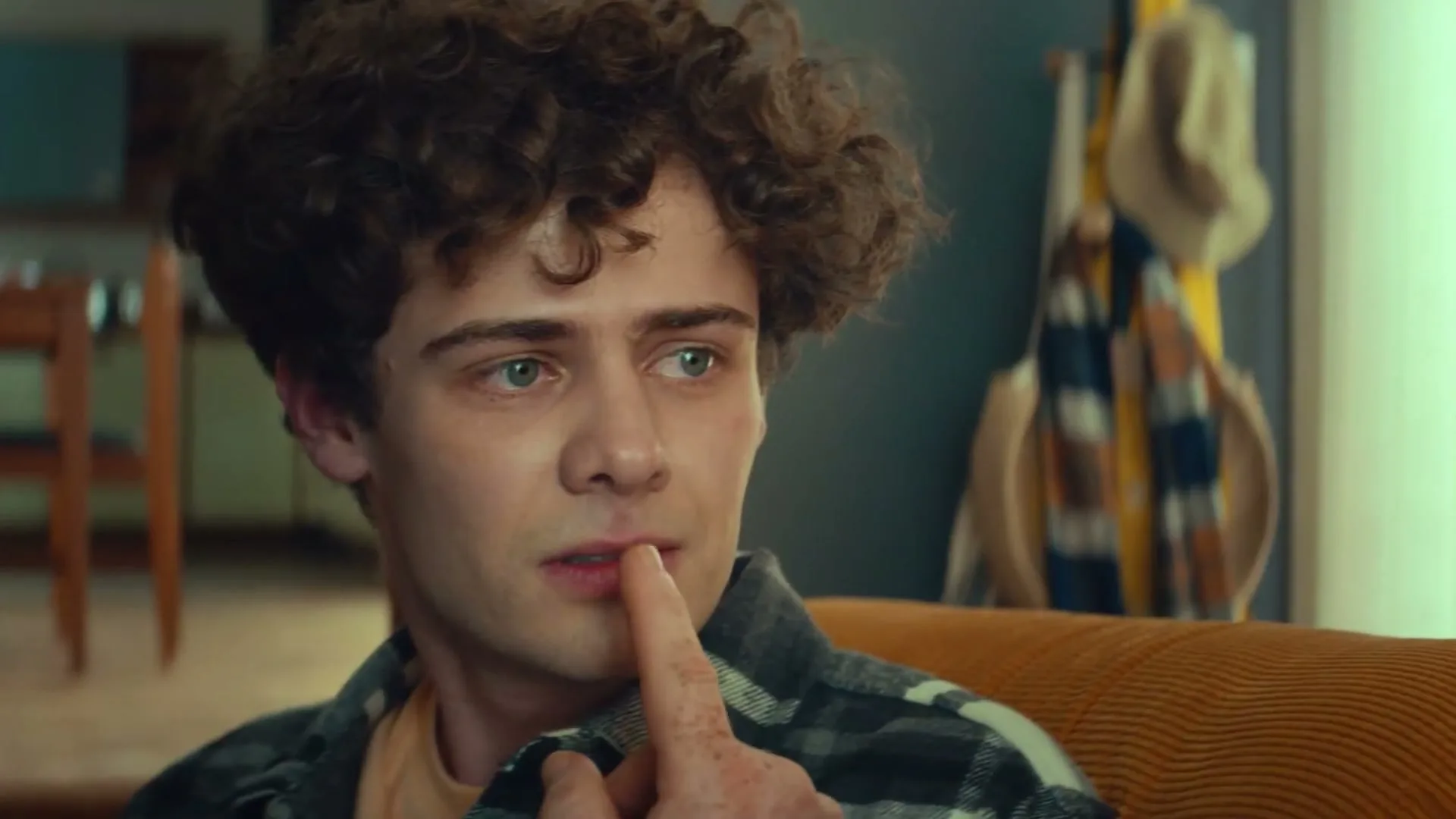 A young man with curly hair sits on an orange couch, looking thoughtful as he touches his nose and then smiles.