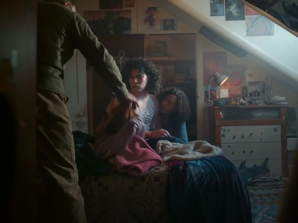 A young person with curly hair sits on a bed, looking distressed, while an older person comforts them in a cluttered attic bedroom.