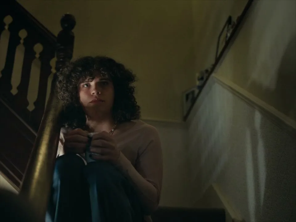A young person with curly hair sits on a dark wooden staircase, looking distressed while holding a small object in their hands.