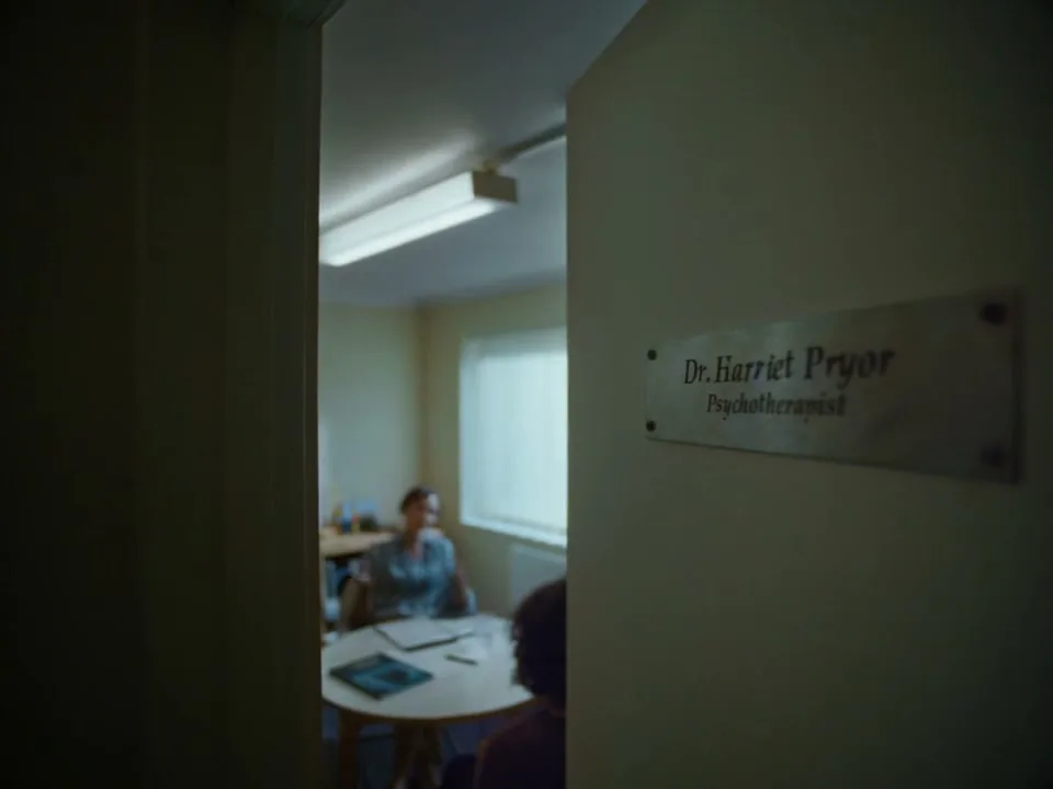 A metal plaque on a door reads "Dr. Harriet Pryor Psychotherapist" while a blurred woman sits at a table in a bright room beyond.