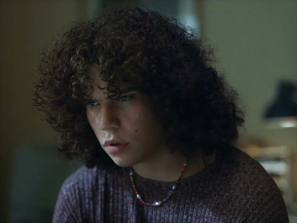 A young person with curly hair and a beaded necklace looks down with a somber expression in an interior setting.
