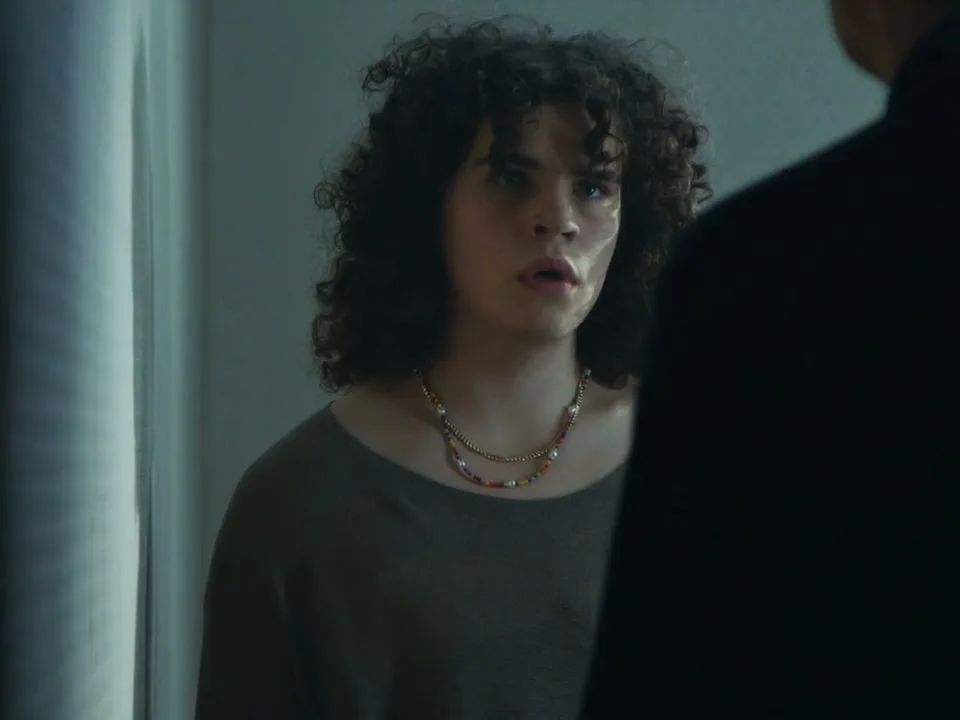 A young person with curly hair looks distressed as they are forced to remove their shirt by an unseen figure in a dimly lit room.