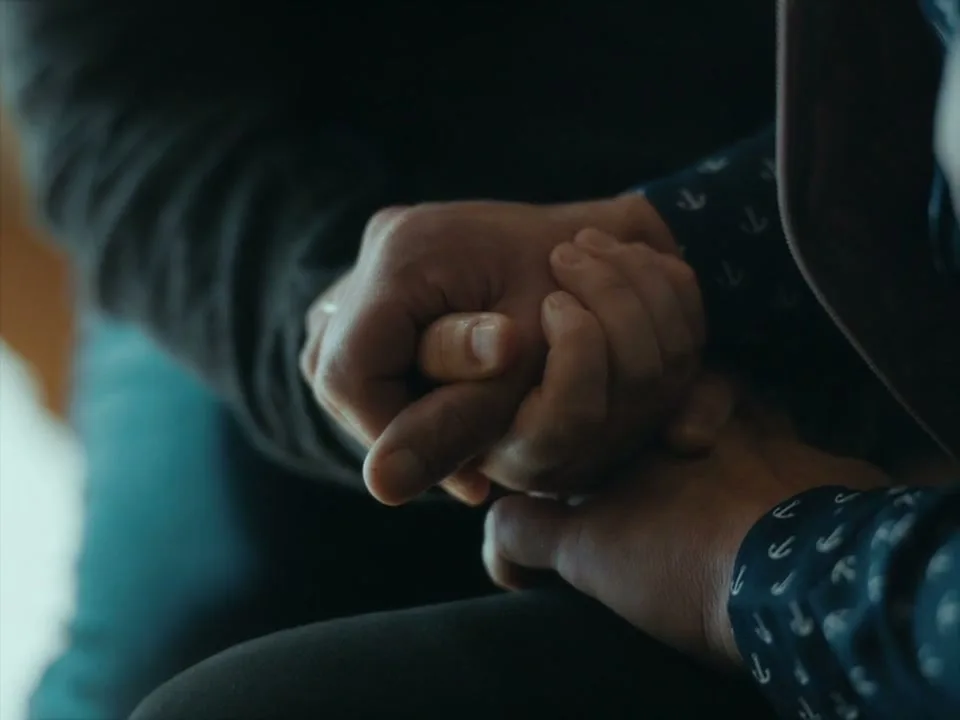 Two people, one wearing a dark long-sleeved shirt and the other a dark blue shirt with white anchors, hold hands tightly in a close-up shot.