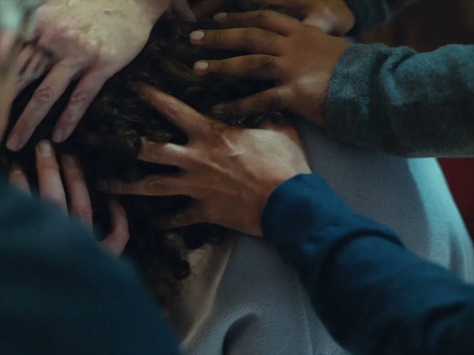 Multiple hands of various skin tones are placed on the head of a person with curly hair, suggesting a moment of intense focus or shared burden in an interior setting.